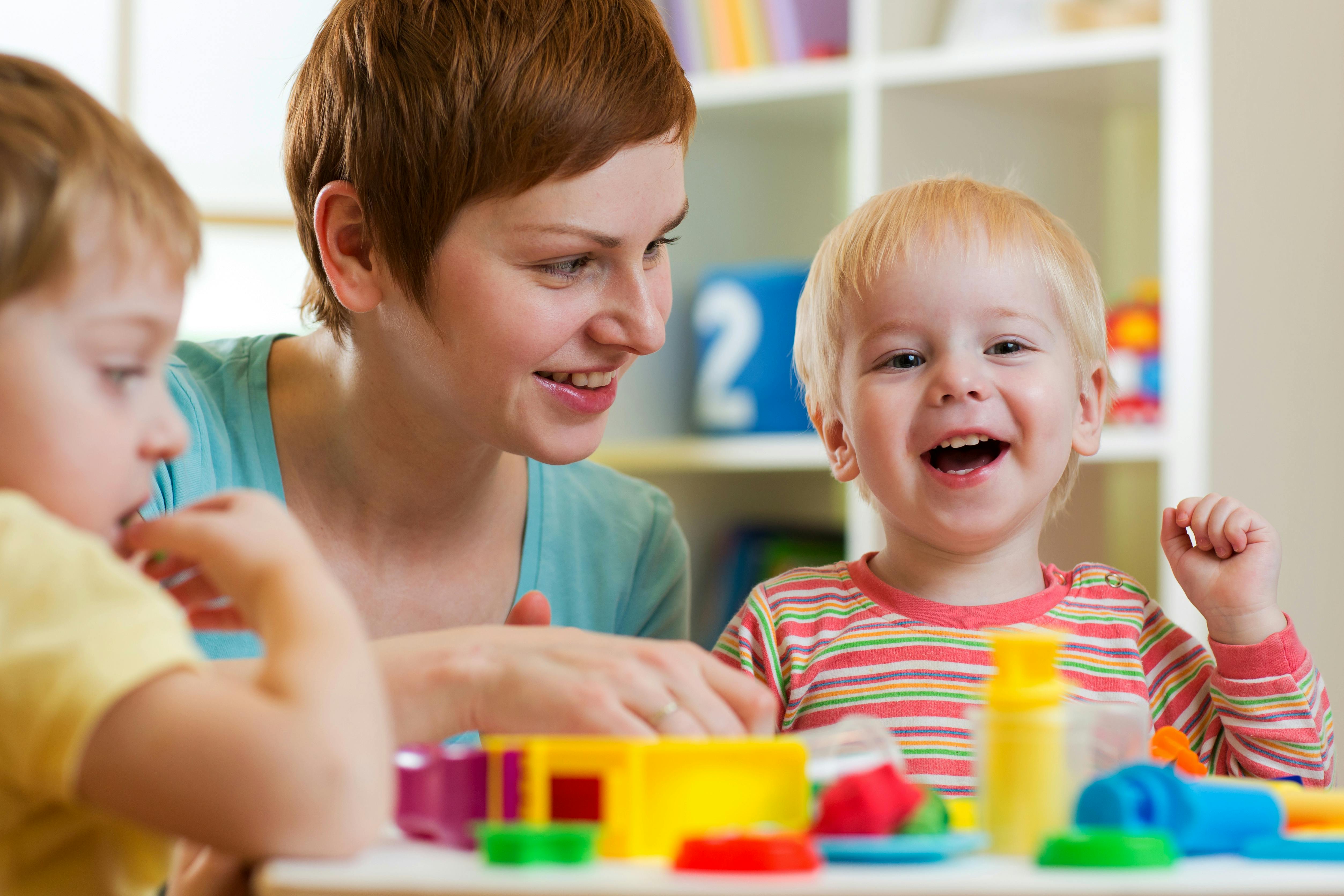 Une femme et deux enfants jouent joyeusement avec des jouets éducatifs colorés dans une salle lumineuse qui ressemble à un espace d'apprentissage pour enfants.