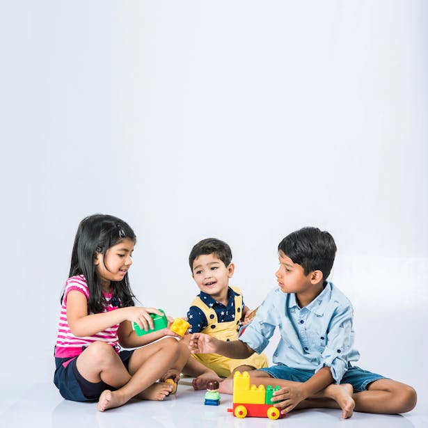 Trois enfants souriants s'assoient ensemble sur le sol et jouent avec des jouets en forme de blocs et de train sur un fond blanc uni.