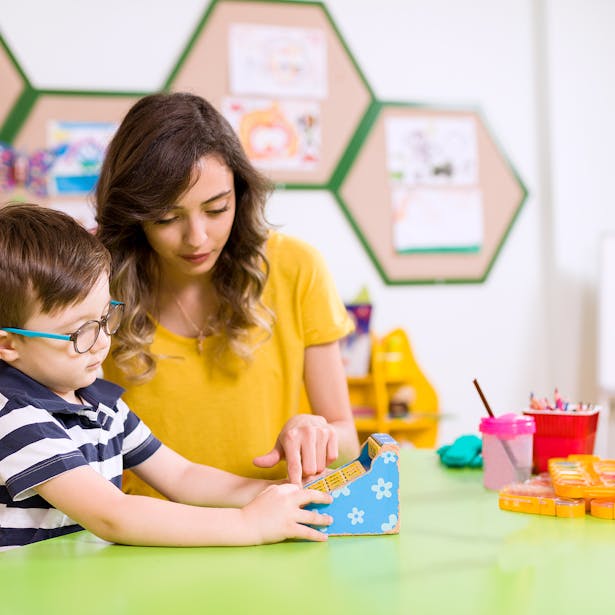Une babysitter aidant un jeune enfant avec des lunettes à faire une activité éducative dans une salle de classe colorée et bien organisée.
