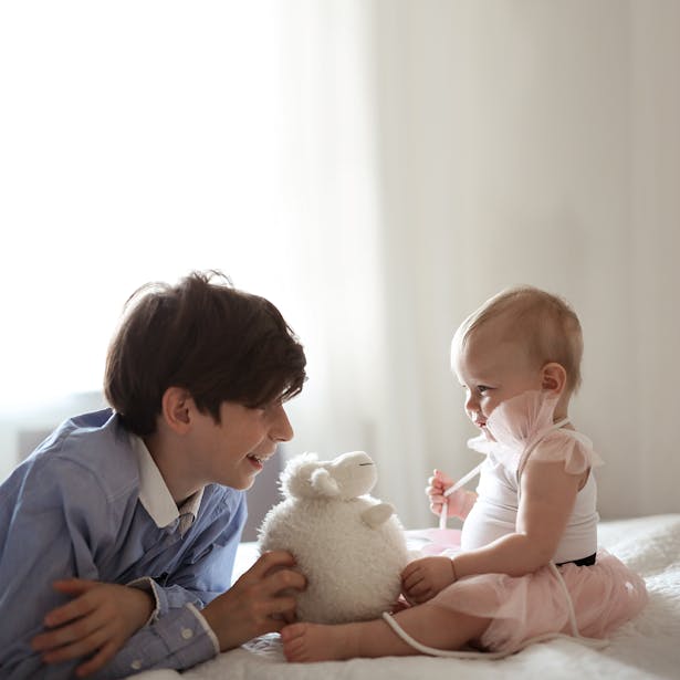 Un jeune garçon jouant avec un bébé qui tient une peluche de mouton, tous deux assis sur un lit dans une pièce éclairée par la lumière naturelle.