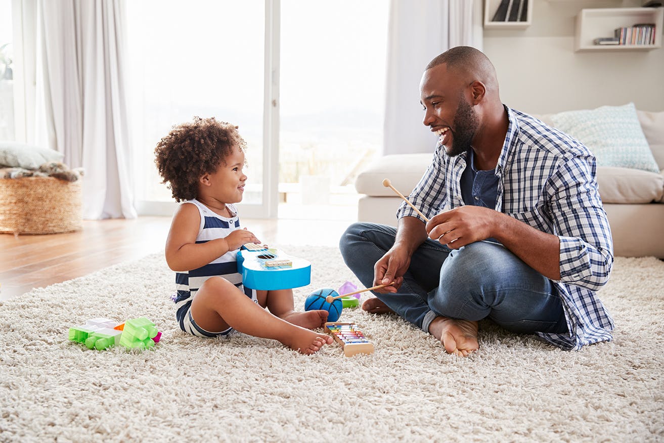 Un babysitter joue avec un enfant, en utilisant des instruments de musique jouets sur un tapis dans un salon ensoleillé.