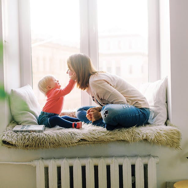Une femme et un bébé jouent et se touchent le nez tendrement, assis sur un banc près d'une fenêtre inondée de lumière naturelle.