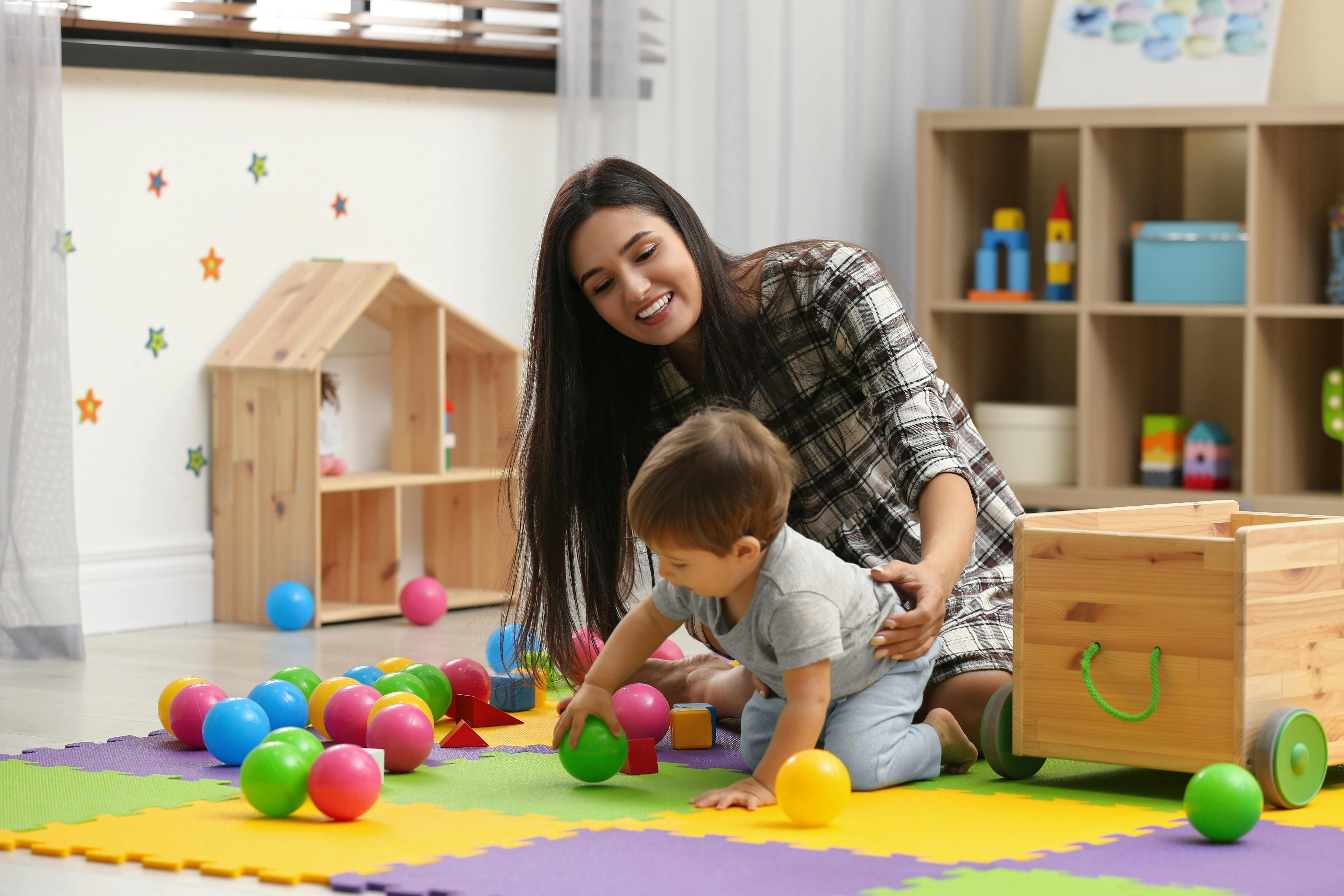 Dans une crèche, une assistante maternelle joue avec un petit garçon par terre avec des boules colorées.