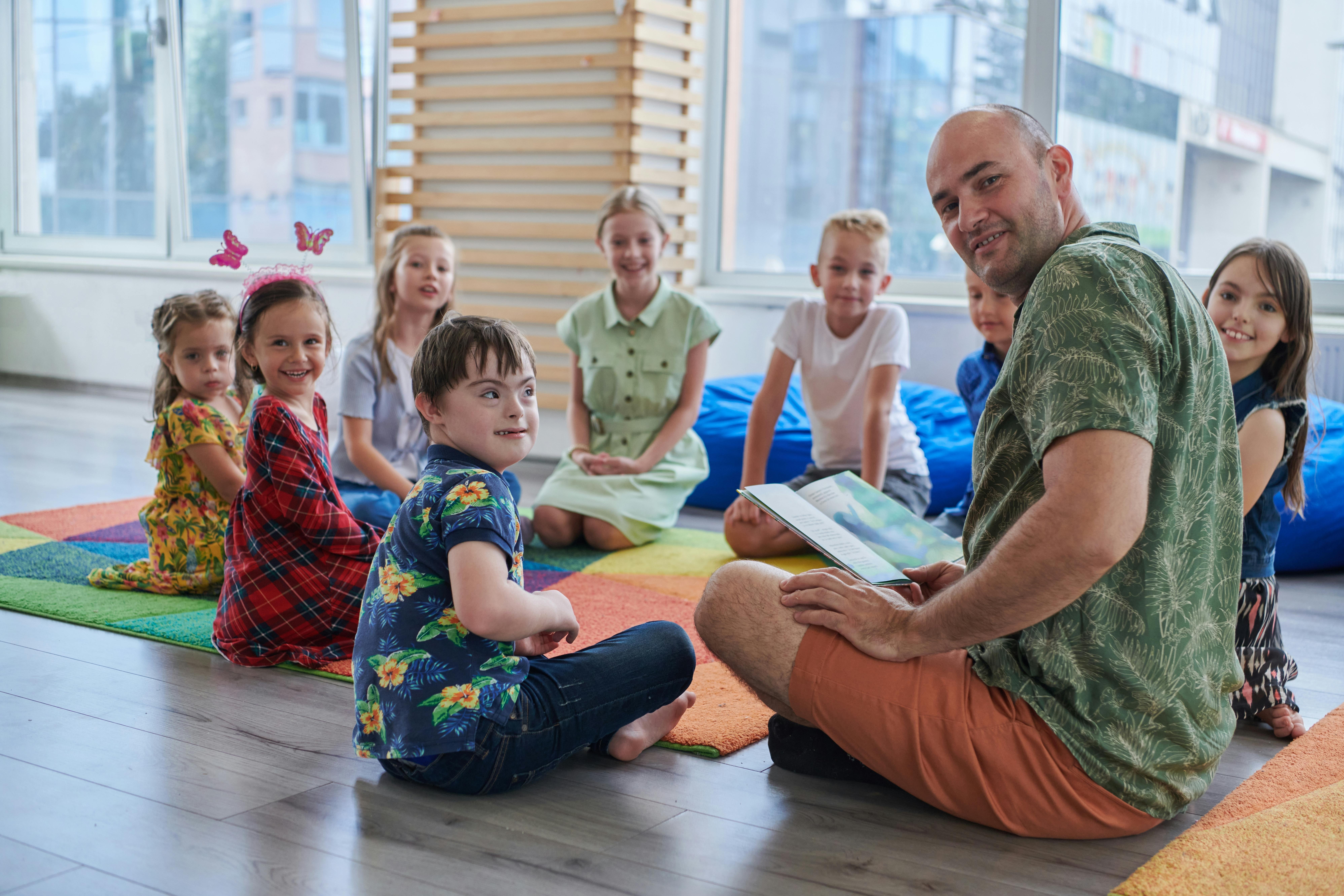 Un groupe d'enfants assis en cercle sur un tapis coloré, écoutant un homme leur lire un livre.