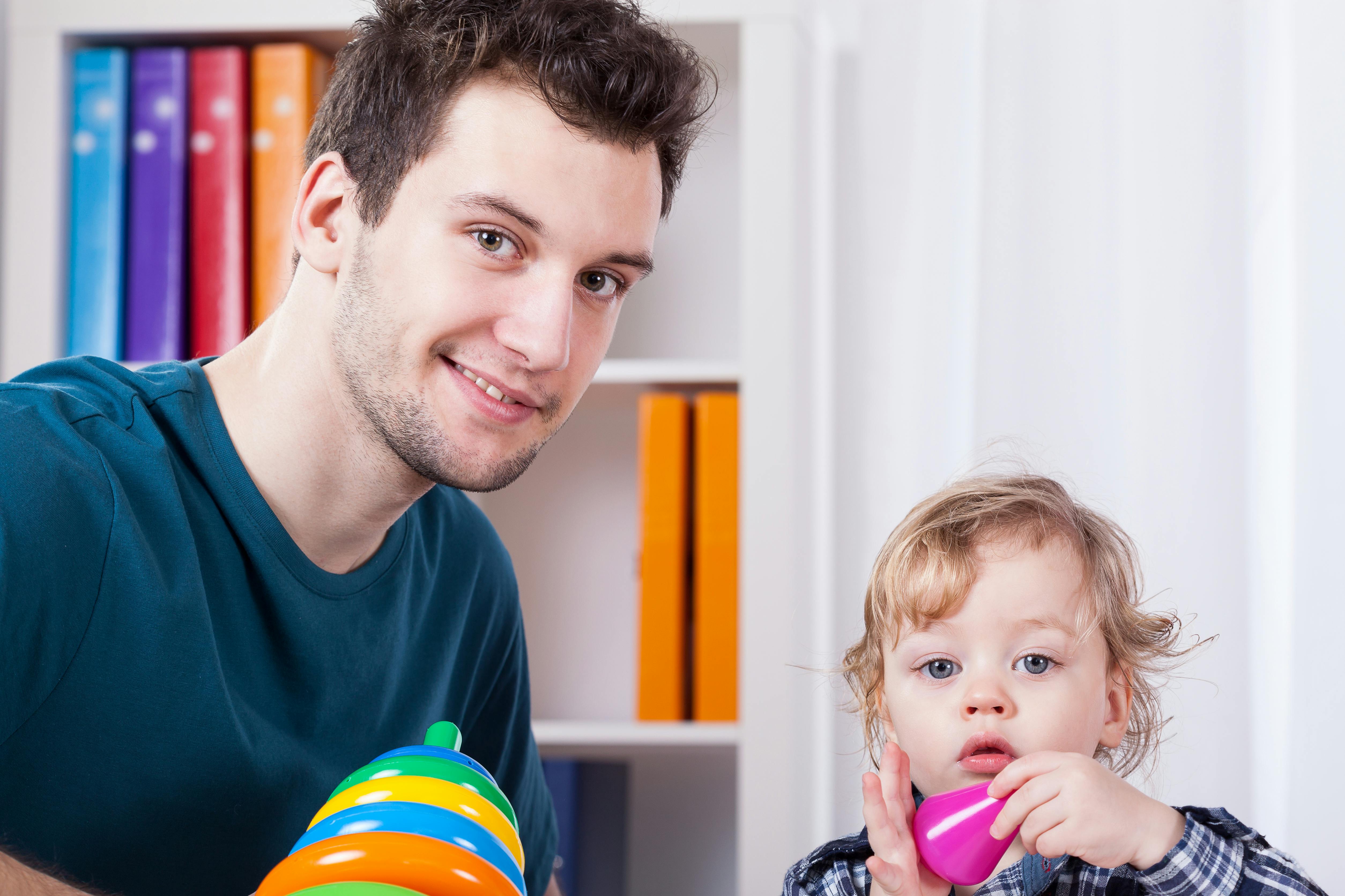Un assistant maternel souriant joue avec son enfant, tenant des jouets colorés.