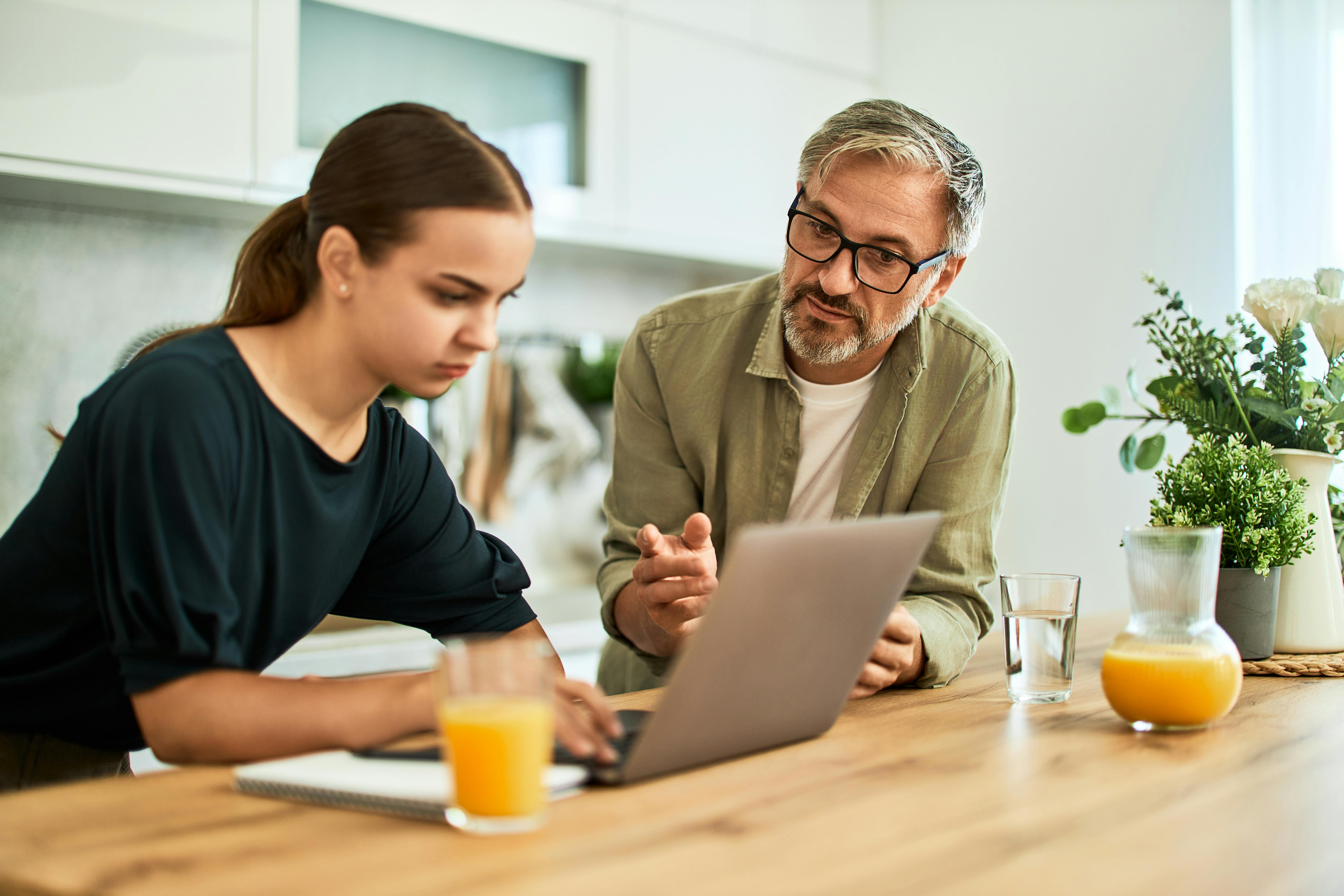 Un homme plus âgé expliquant quelque chose à une jeune femme concentrée sur un ordinateur portable à une table de cuisine.