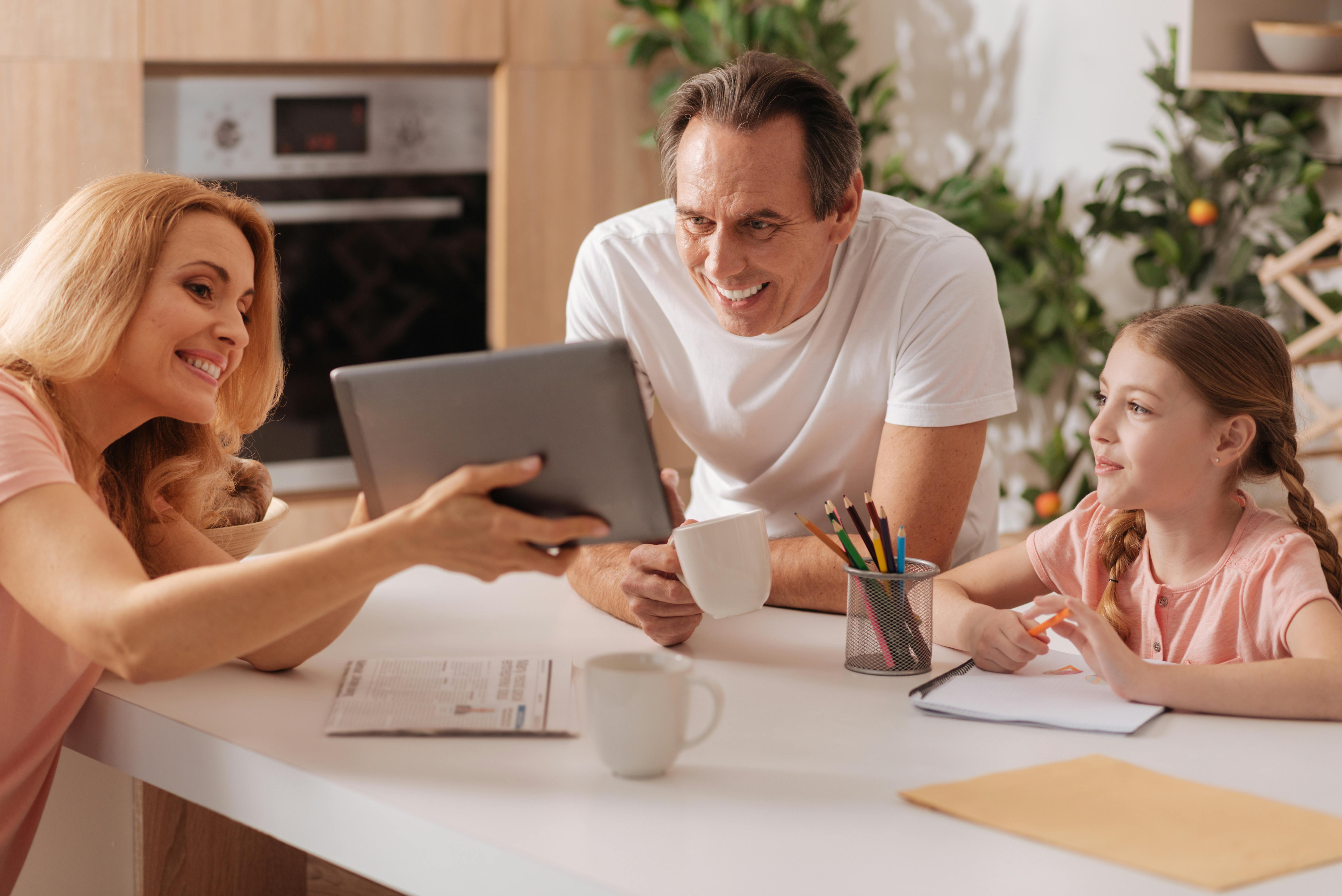 Une famille regardant une tablette ensemble à une table de cuisine, souriant et interagissant joyeusement.