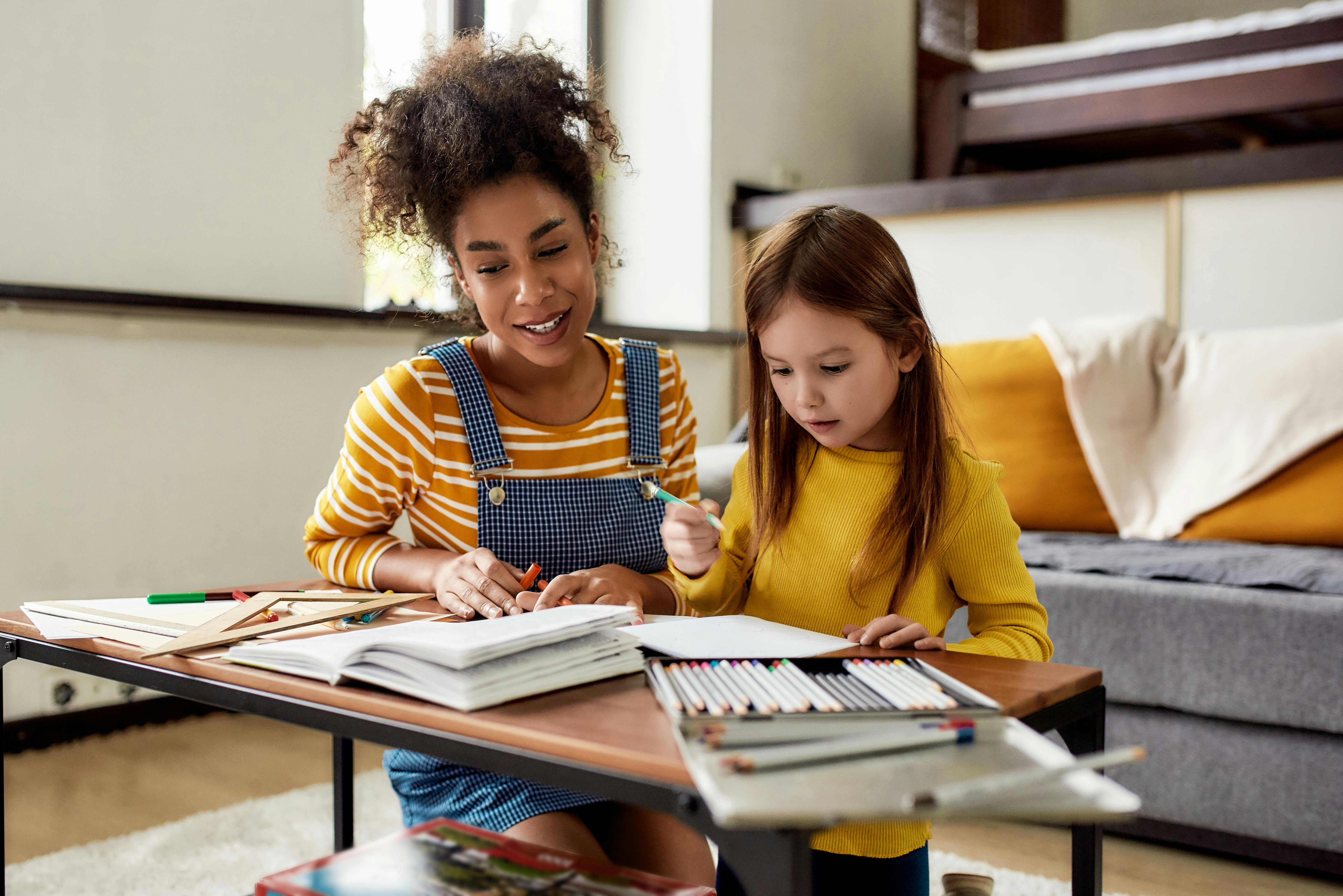 Une enseignante aide une petite fille à dessiner et colorier, assises à une table dans une pièce chaleureuse et accueillante.