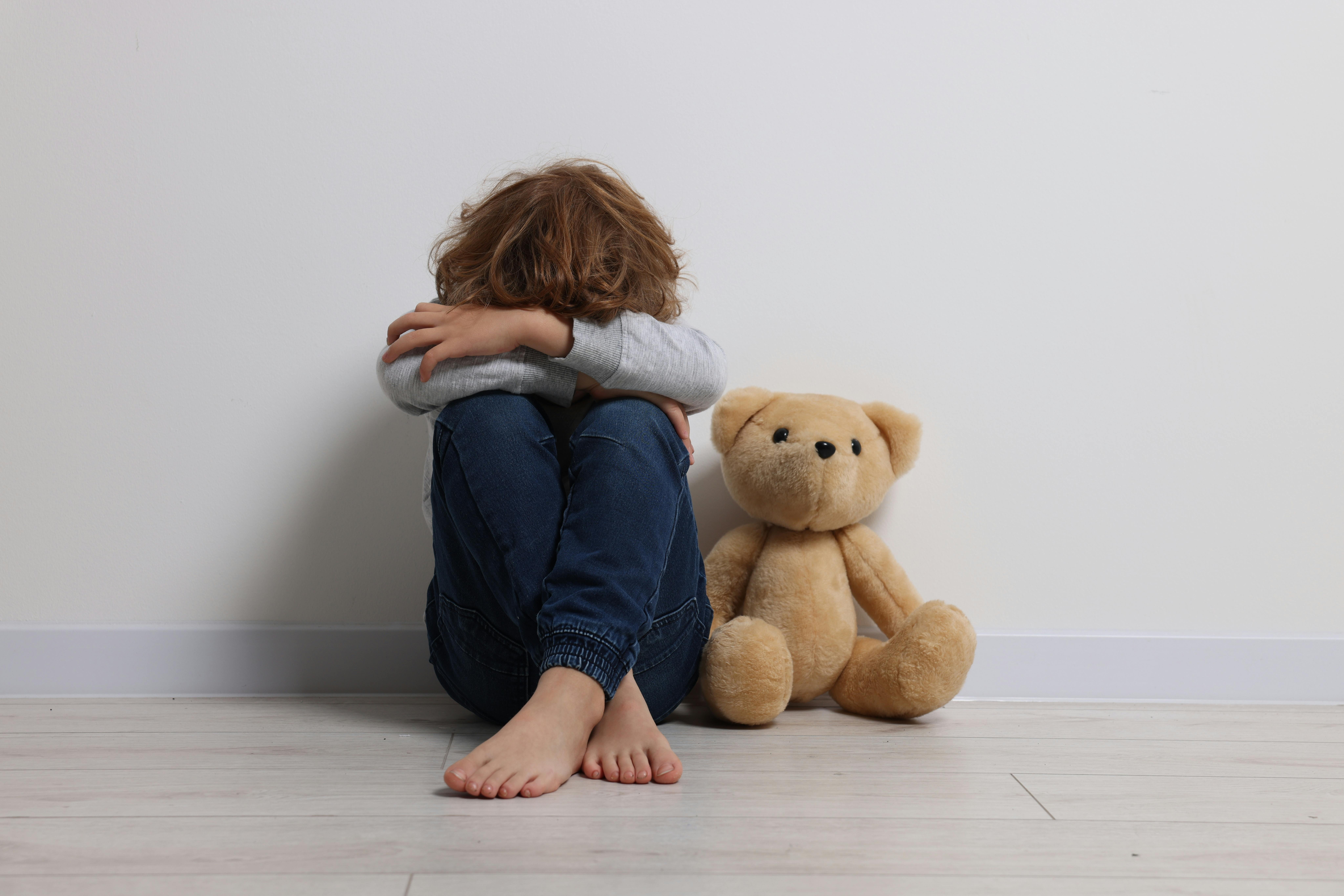 Un enfant semble triste et réconforté par la présence d'un ours en peluche à ses côtés. Assis sur le sol, l'enfant cache son visage dans ses genoux, créant une atmosphère de solitude ou de réconfort.