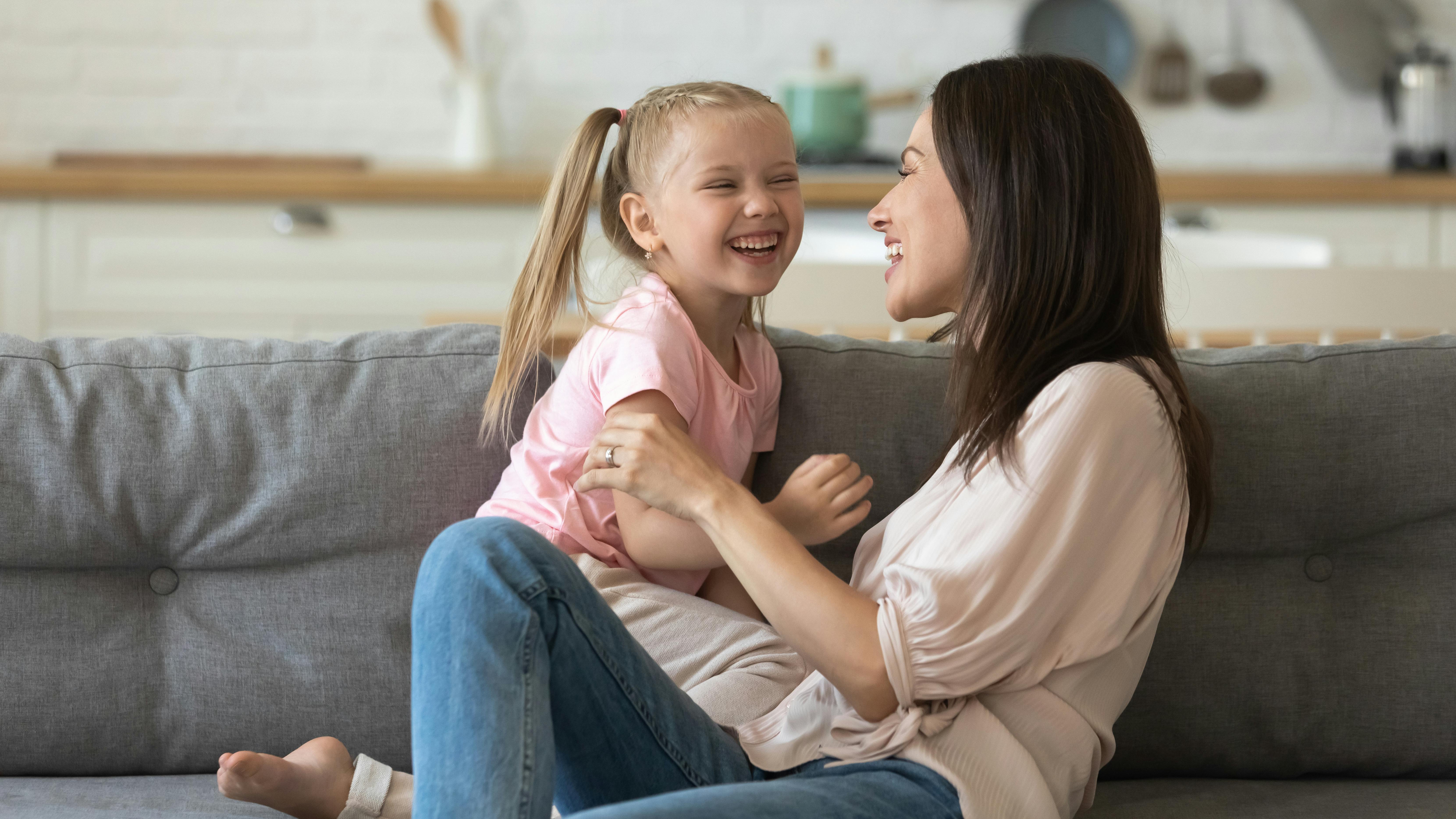 Une mère et sa fille rient ensemble, assises sur un canapé dans une cuisine lumineuse.