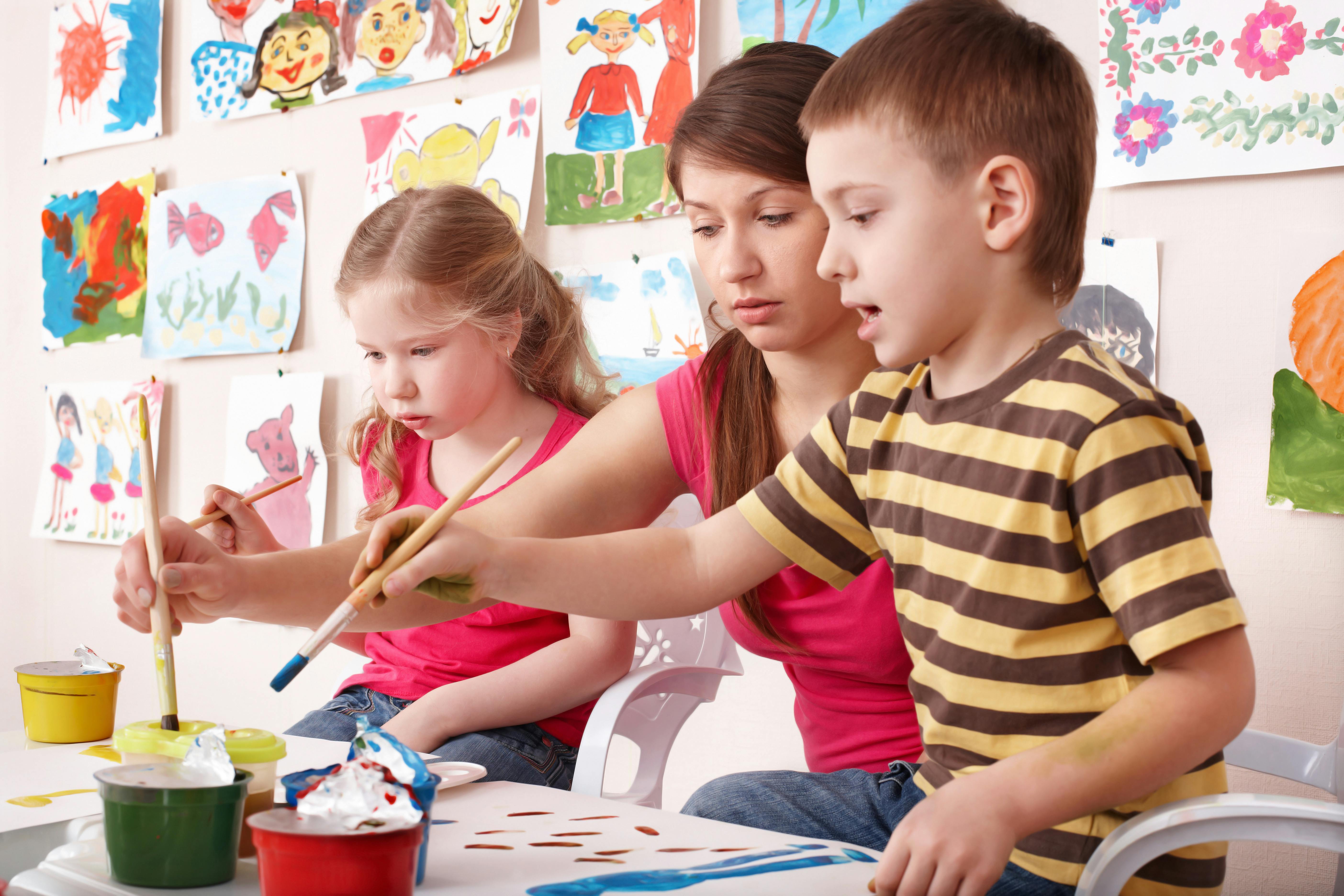 Deux enfants peignant avec des pinceaux, aidés par une femme, dans une salle décorée avec des dessins d'enfants accrochés au mur.