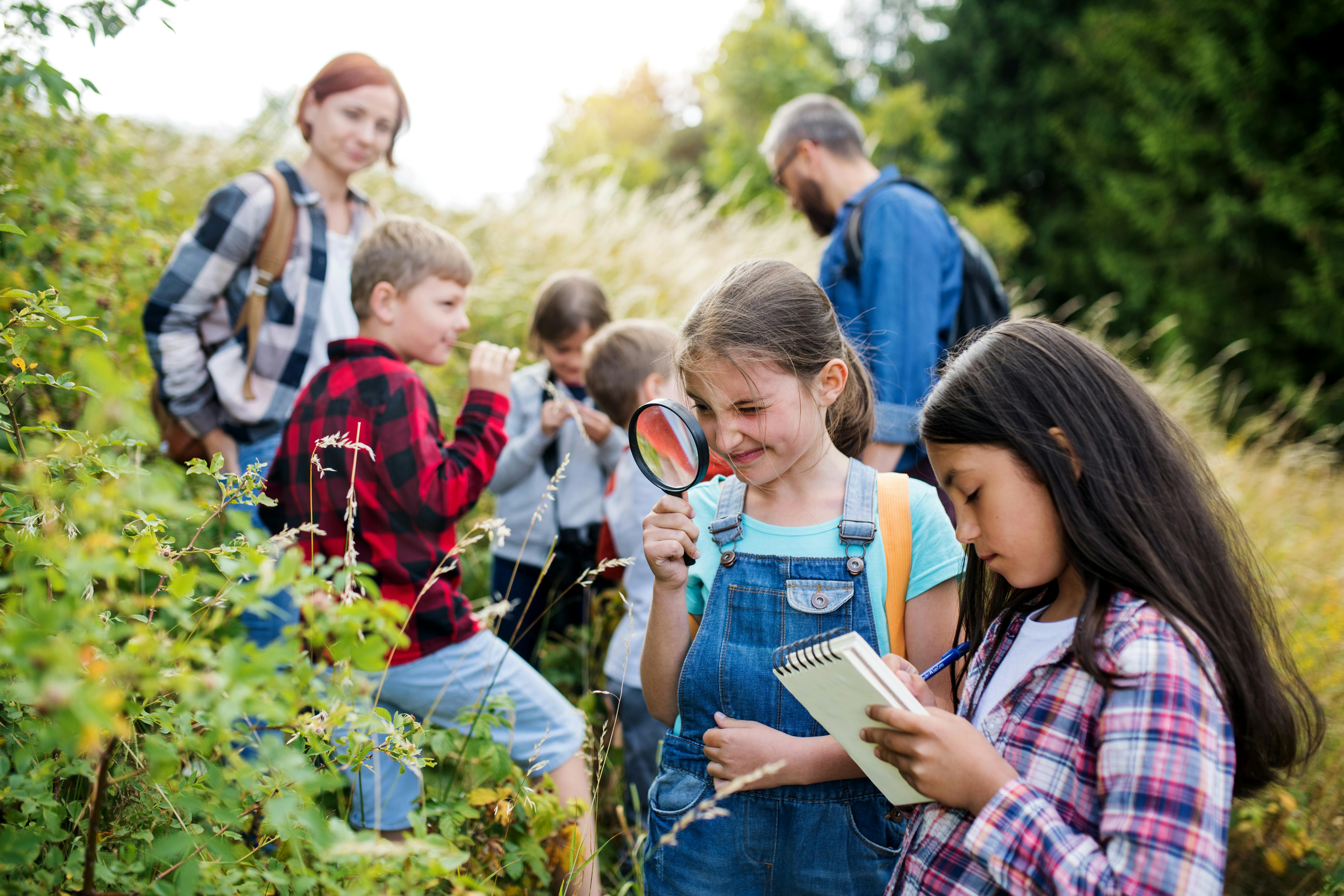 Un groupe d'enfants explore la nature avec des loupes et des carnets, accompagnés de deux adultes.