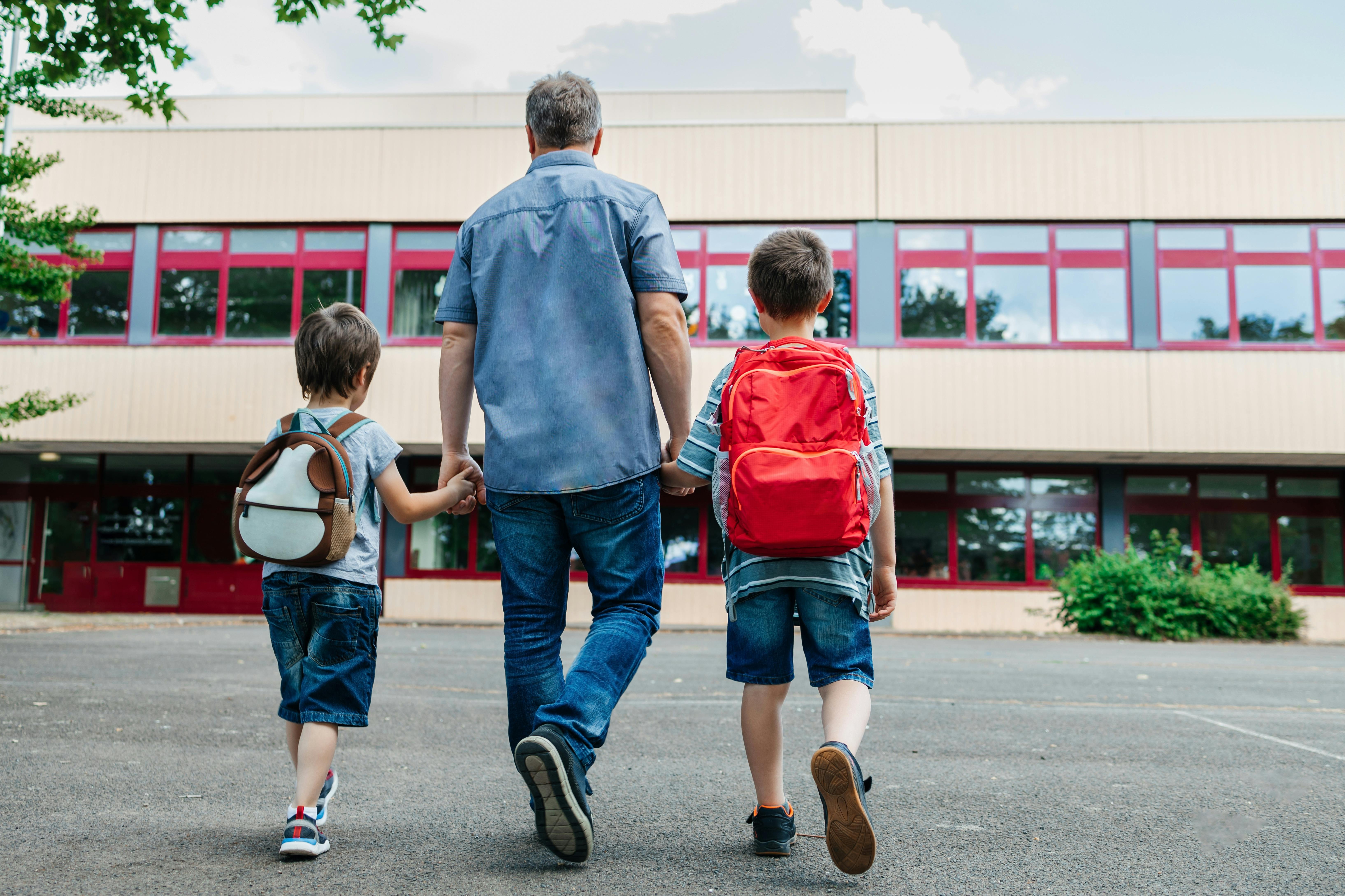 Un père accompagne ses deux enfants à l'école, vus de dos.