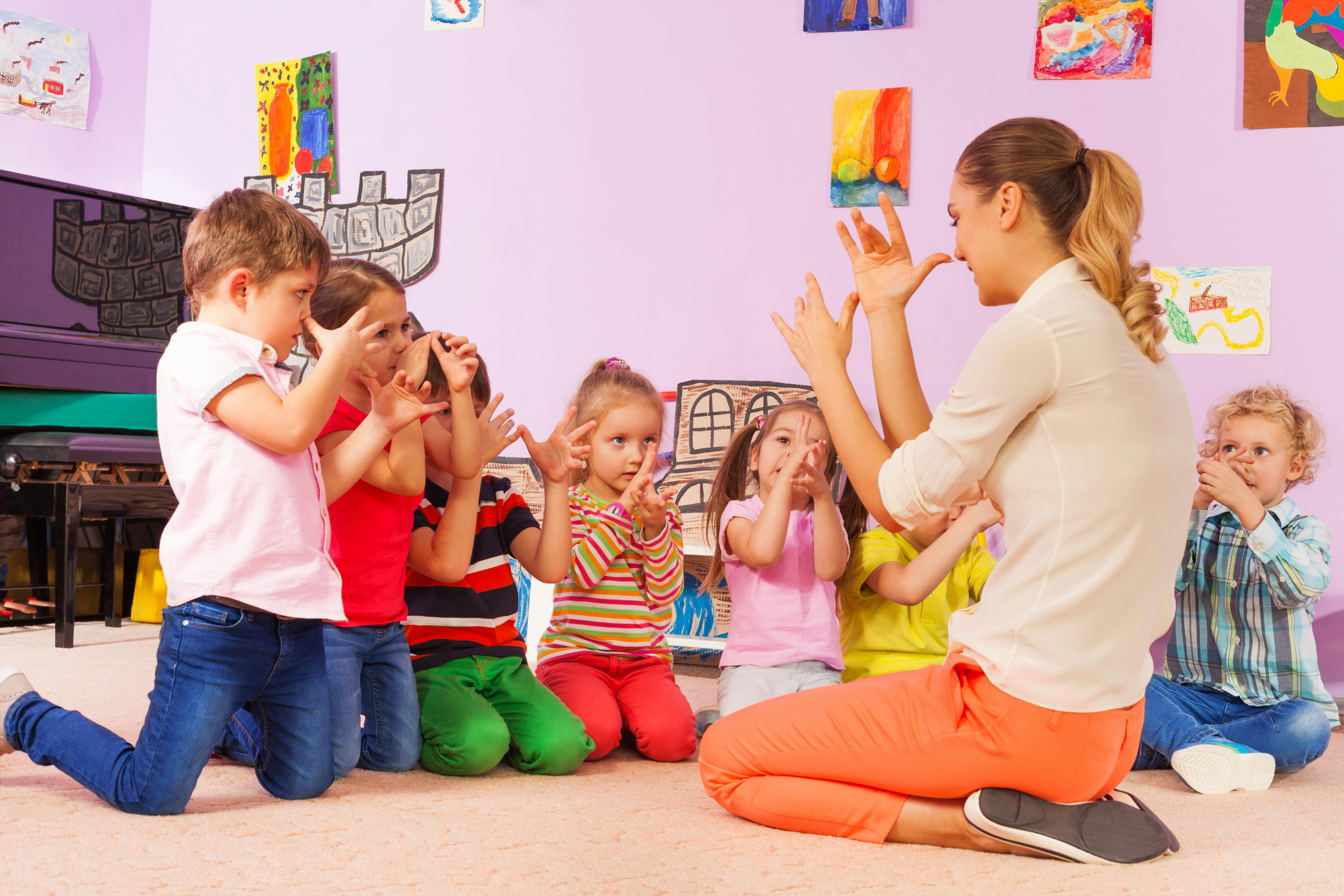 Une enseignante anime une activité de chant et de gestuelle avec un groupe d'enfants dans une salle de classe décorée.