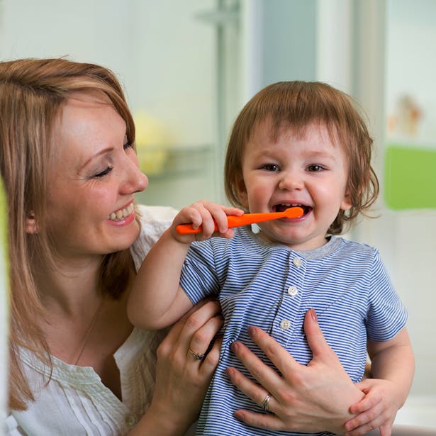 Une femme souriante tenant un jeune enfant qui s'amuse à se brosser les dents avec une brosse à dents orange dans une salle de bain lumineuse.