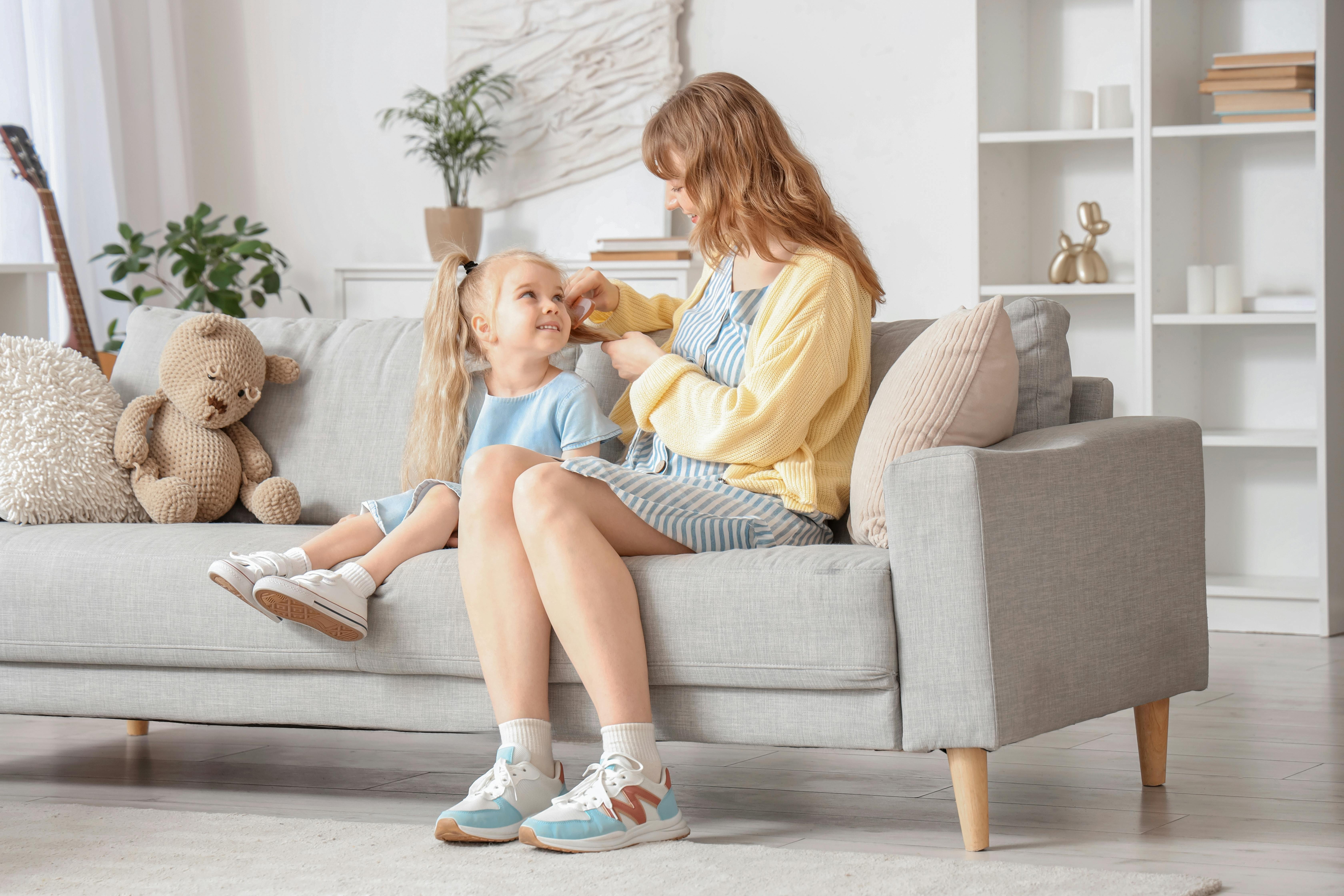 Une jeune babysitter est en train de brosser les cheveux d'une petite fille. Les deux sont assises sur le canapé du salon.