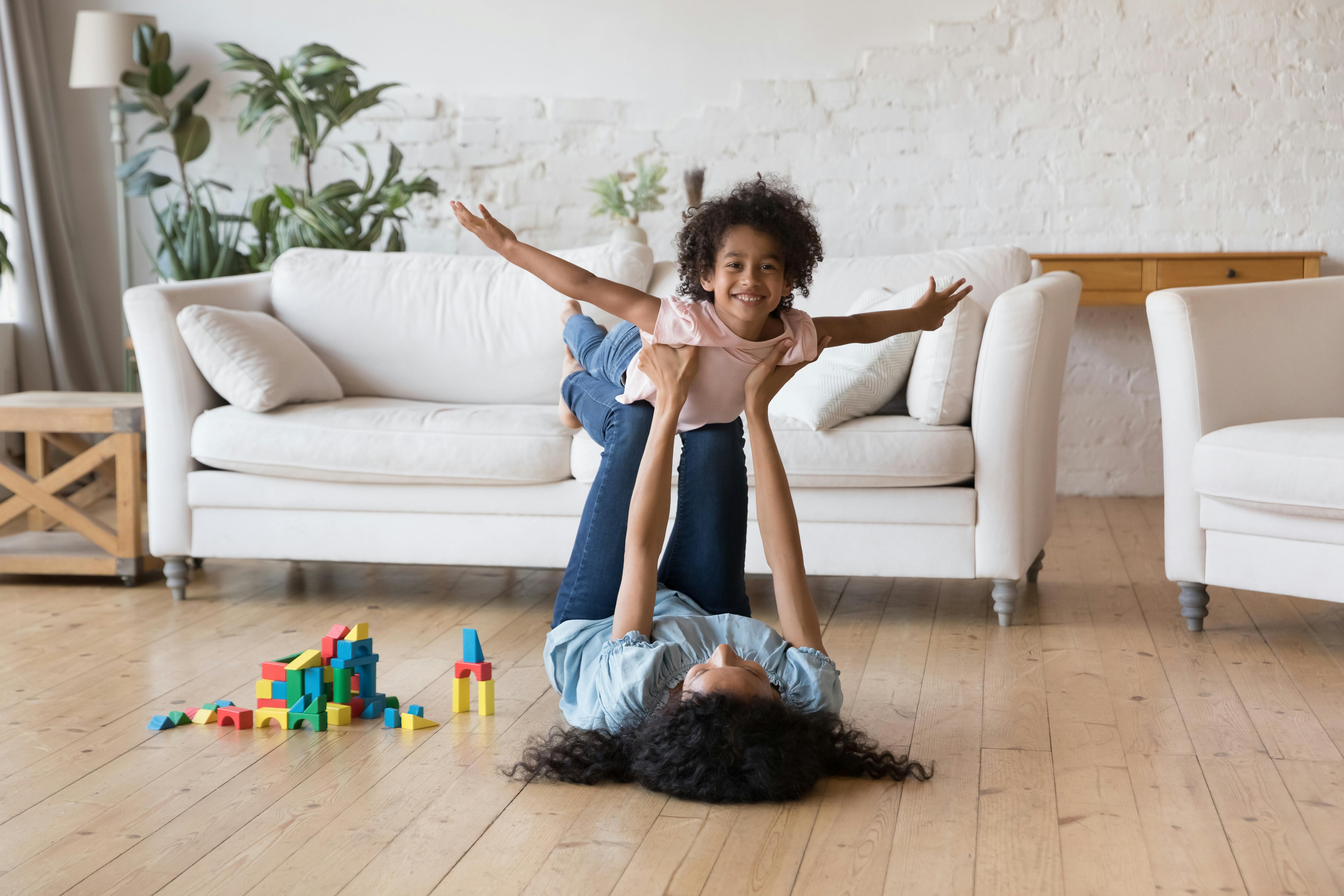 Une babysitter joue avec une petite fille au milieu d'un salon. La jeune fille imite un avion.