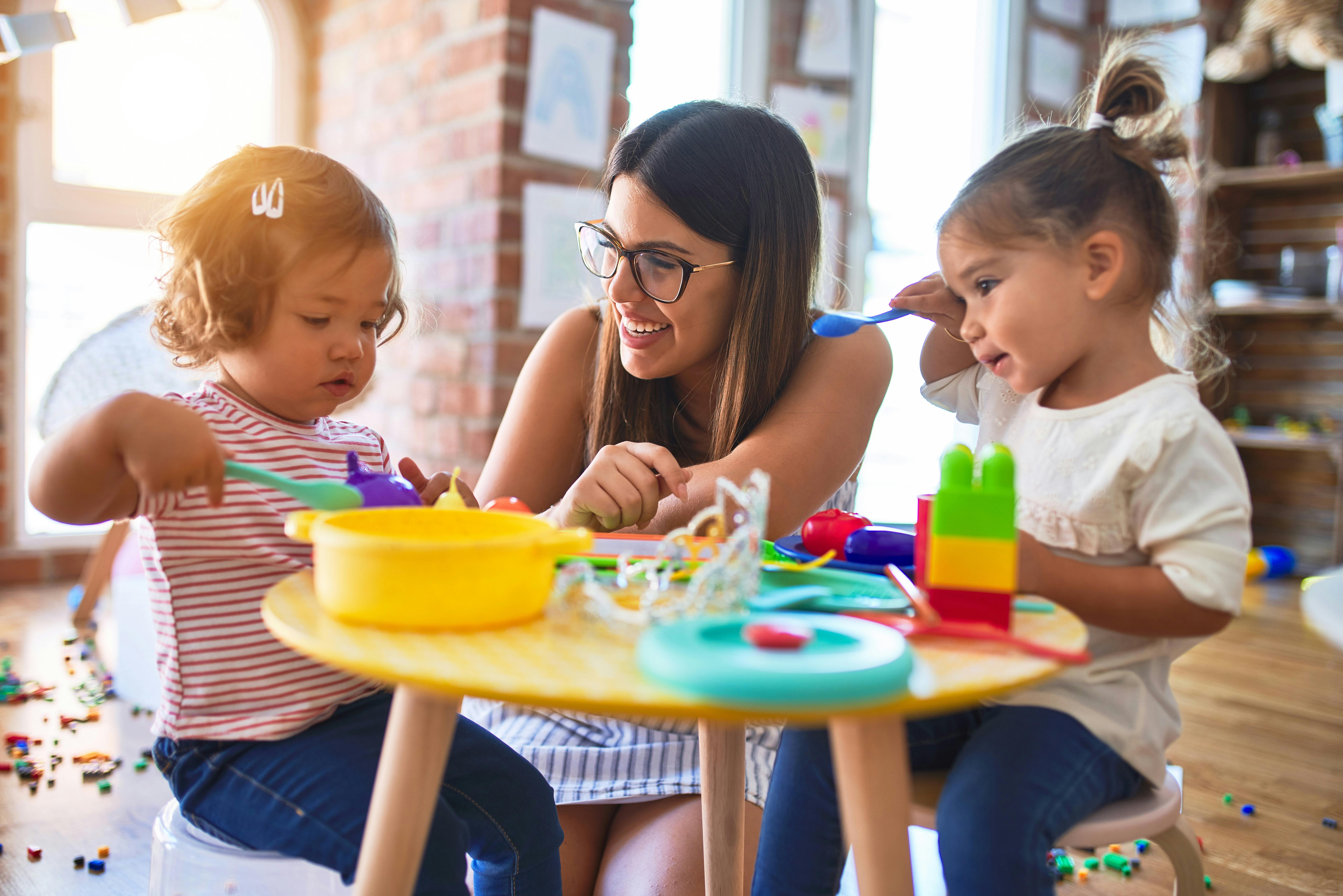 Une éducatrice joue à la dinette avec deux jeunes enfants dans une salle de jeux bien éclairée.