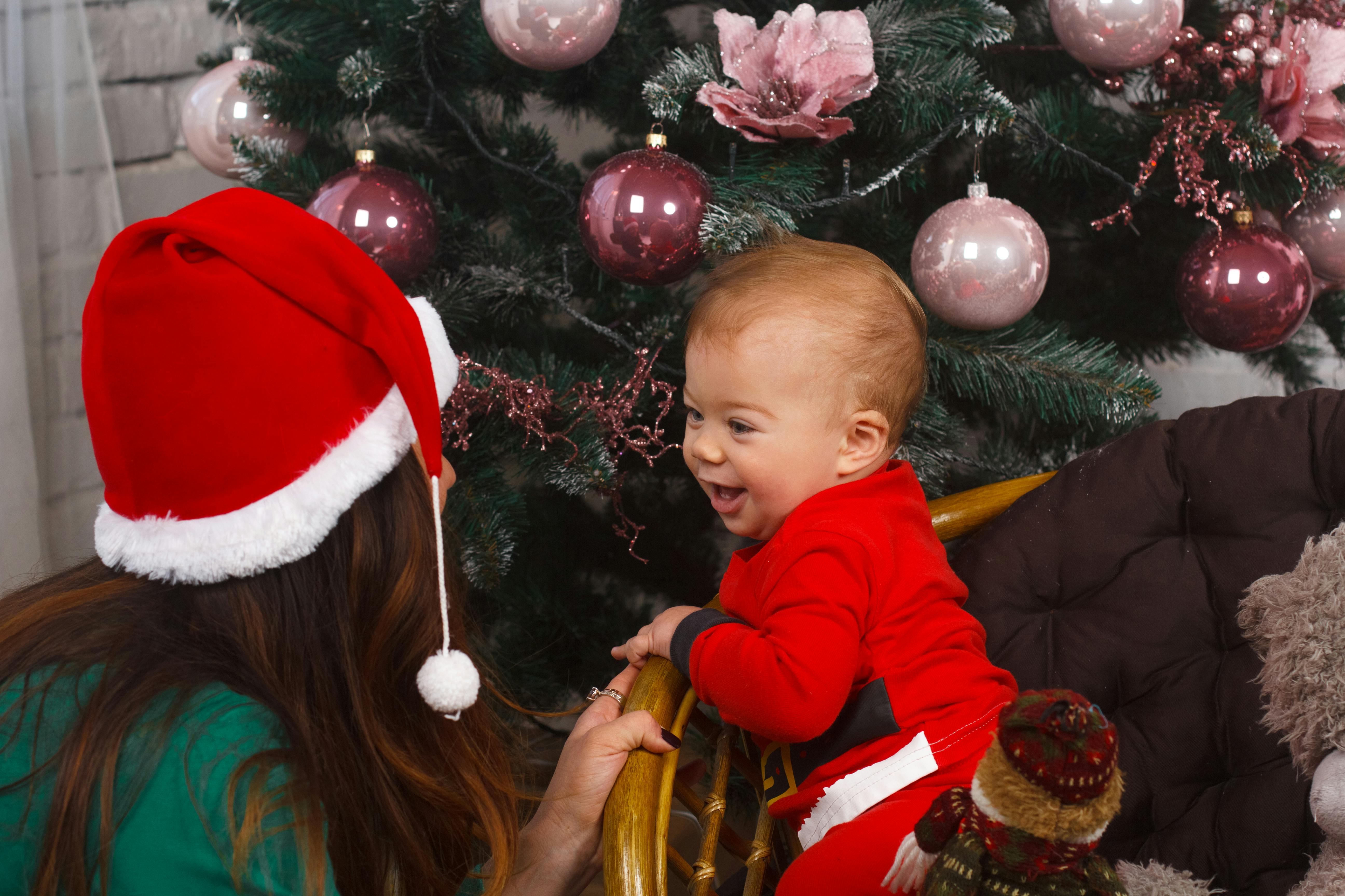 Un bébé déguisé en père Noël est à genoux sur une chaise à côté d'un sapin pendant que sa gardienne le surveille, portant un bonnet de Noël.
