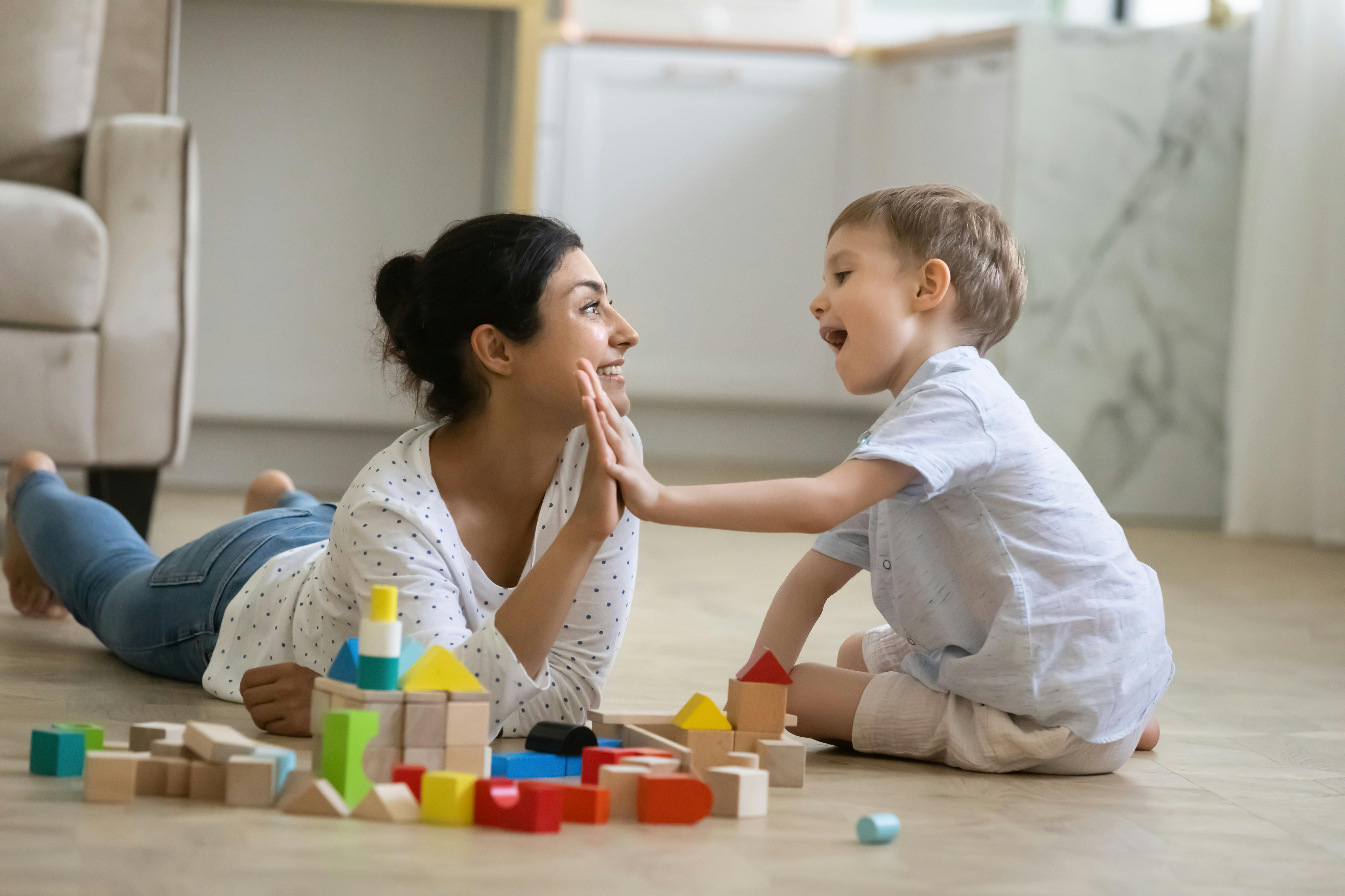 Une jeune assistante maternelle et un petit garçon se font un high five après avoir construit une maison avec des blocs de construction.