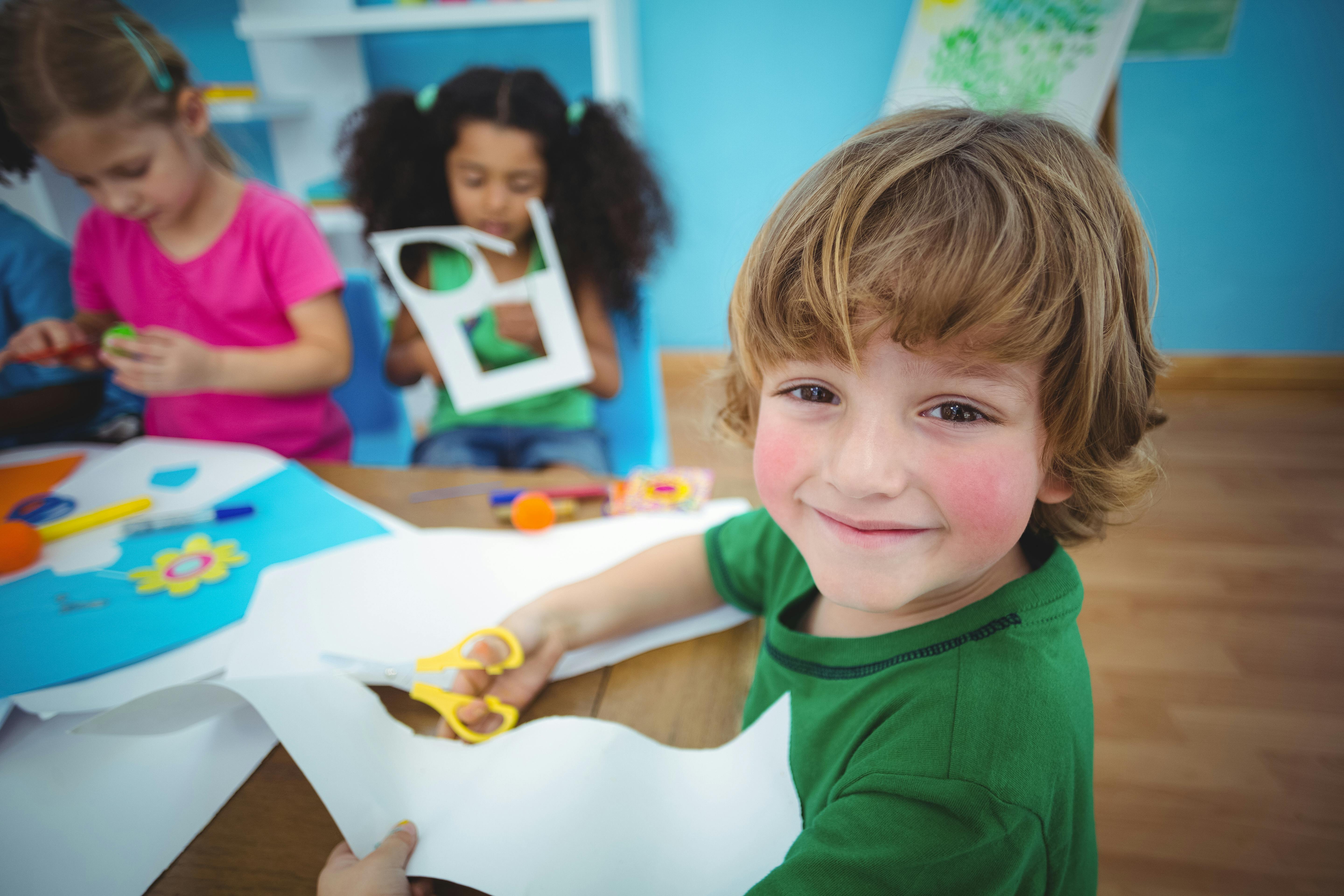 Un petit garçon regarde intensément la caméra en souriant, pendant qu'il fait des activités manuelles au milieu de ses petits camarades.