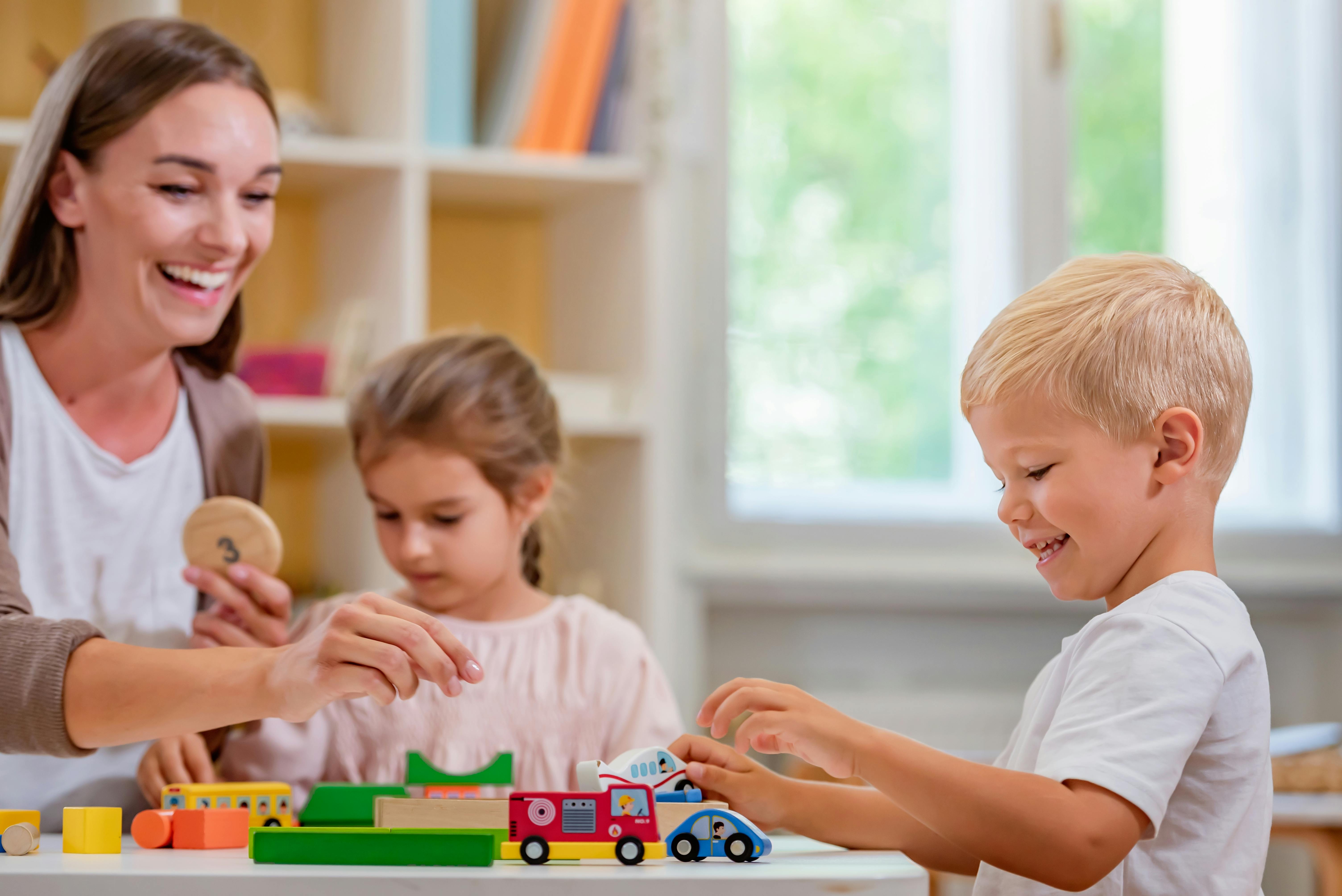 Une enseignante joue avec deux jeunes enfants en utilisant des jouets en bois et des voitures miniatures, créant un environnement d'apprentissage joyeux.