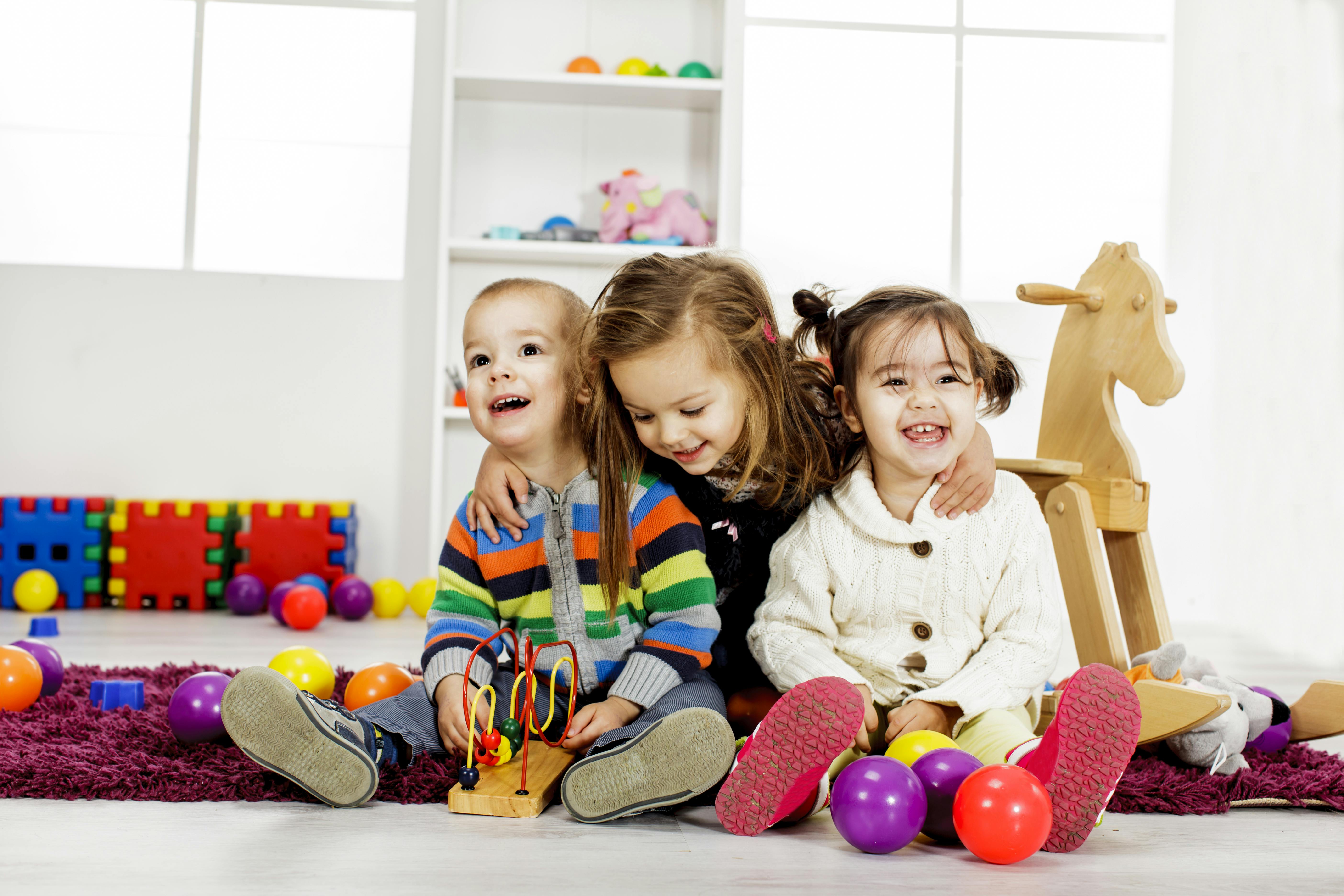 Trois jeunes enfants assis ensemble sur le sol d'une salle de jeux, souriant et riant, avec des jouets colorés autour d'eux.