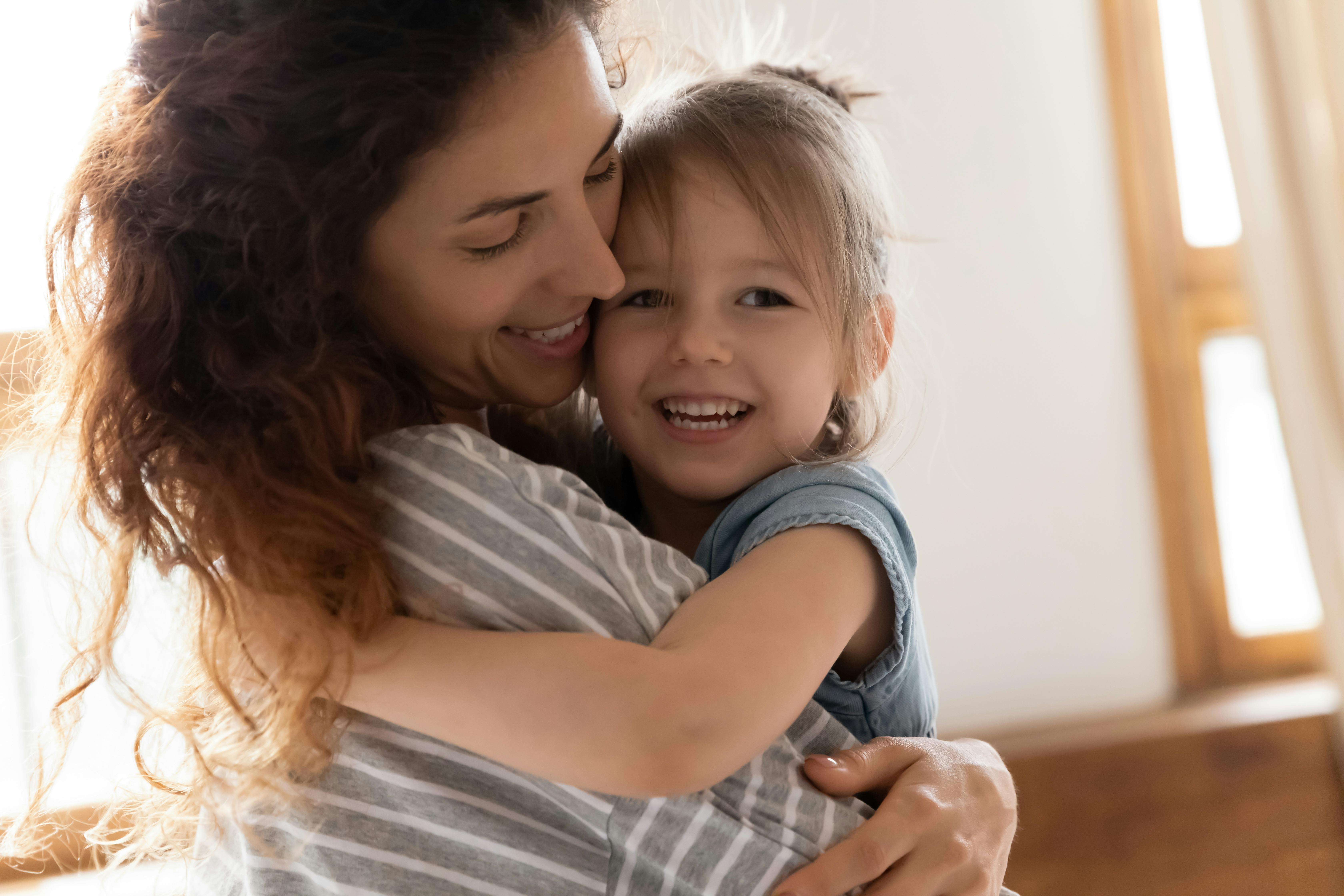 Une mère câline sa fille souriante dans un intérieur lumineux.