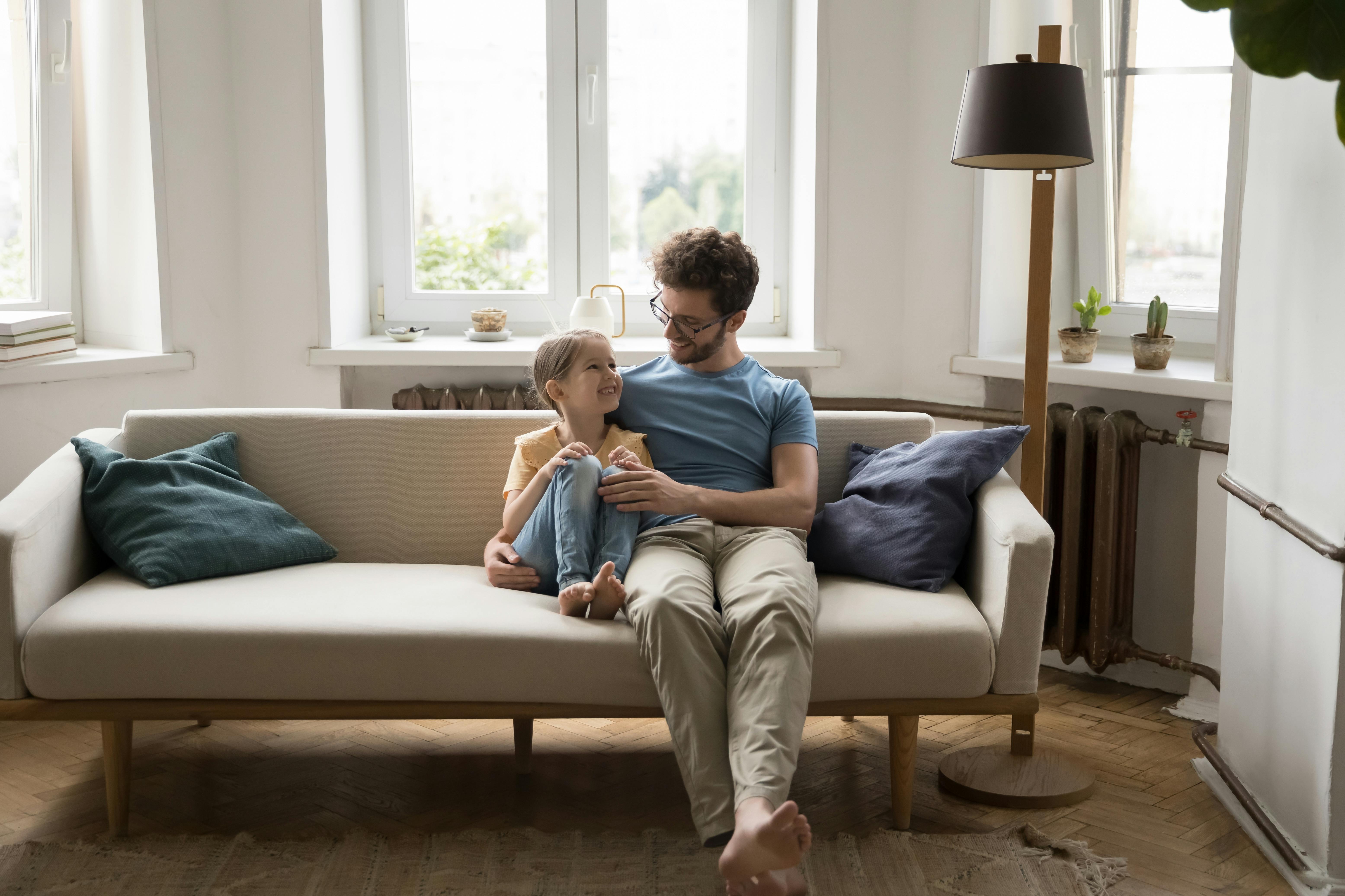 Un père et sa fille rient ensemble sur un canapé dans un salon lumineux.