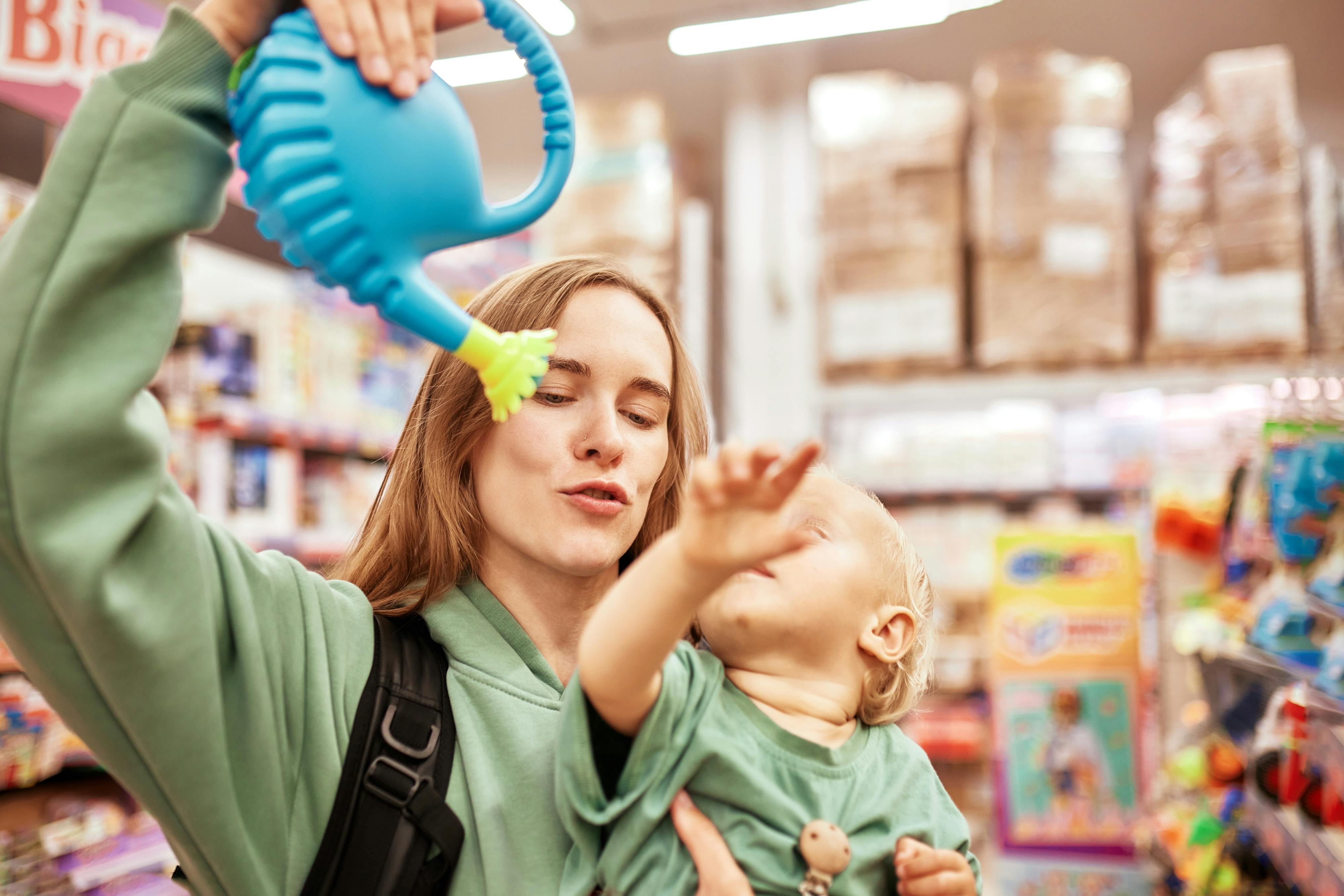 Une femme tient un arrosoir bleu en plastique et un bébé dans un magasin de jouets.