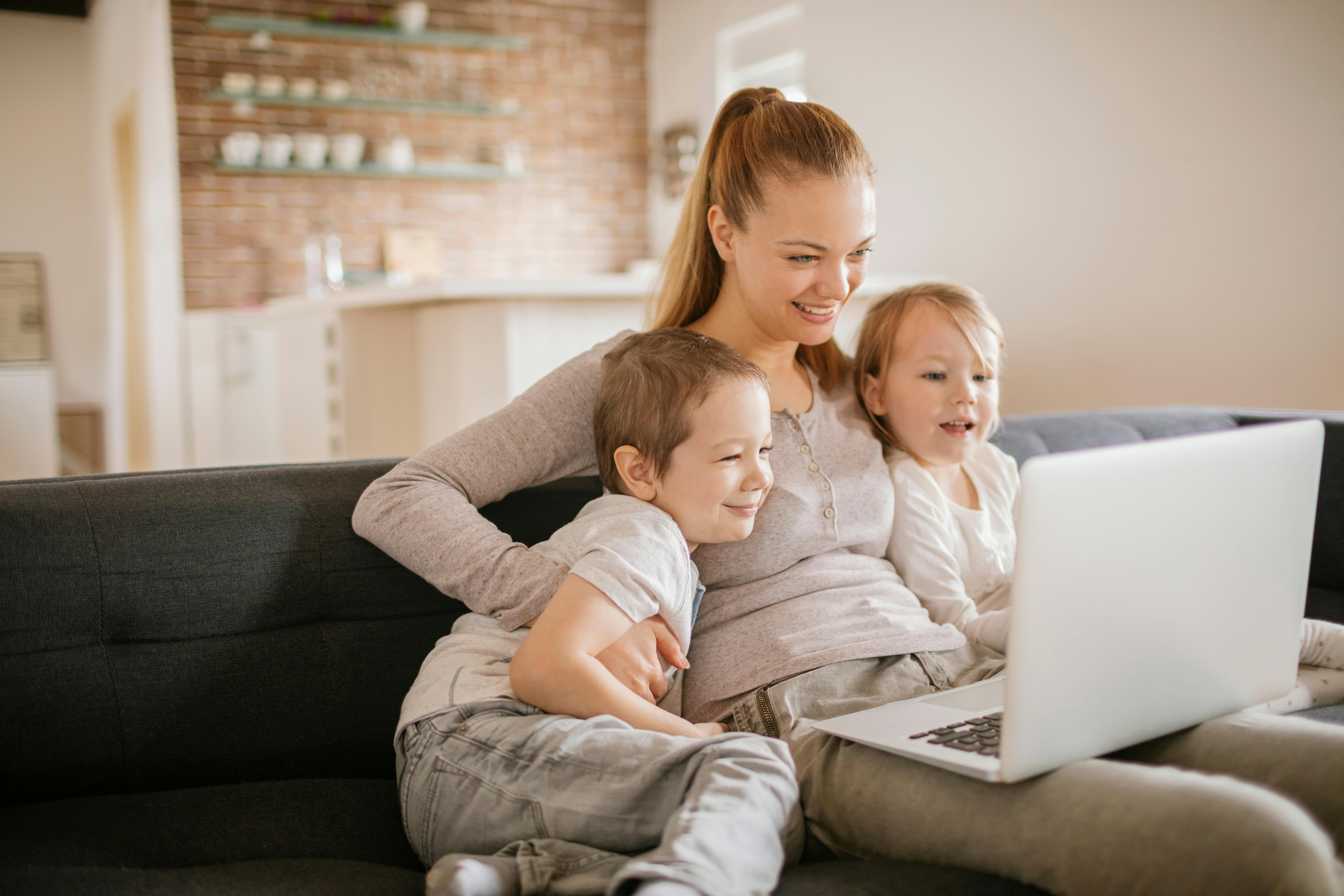 Une femme regarde un ordinateur portable avec deux enfants assis sur un canapé.