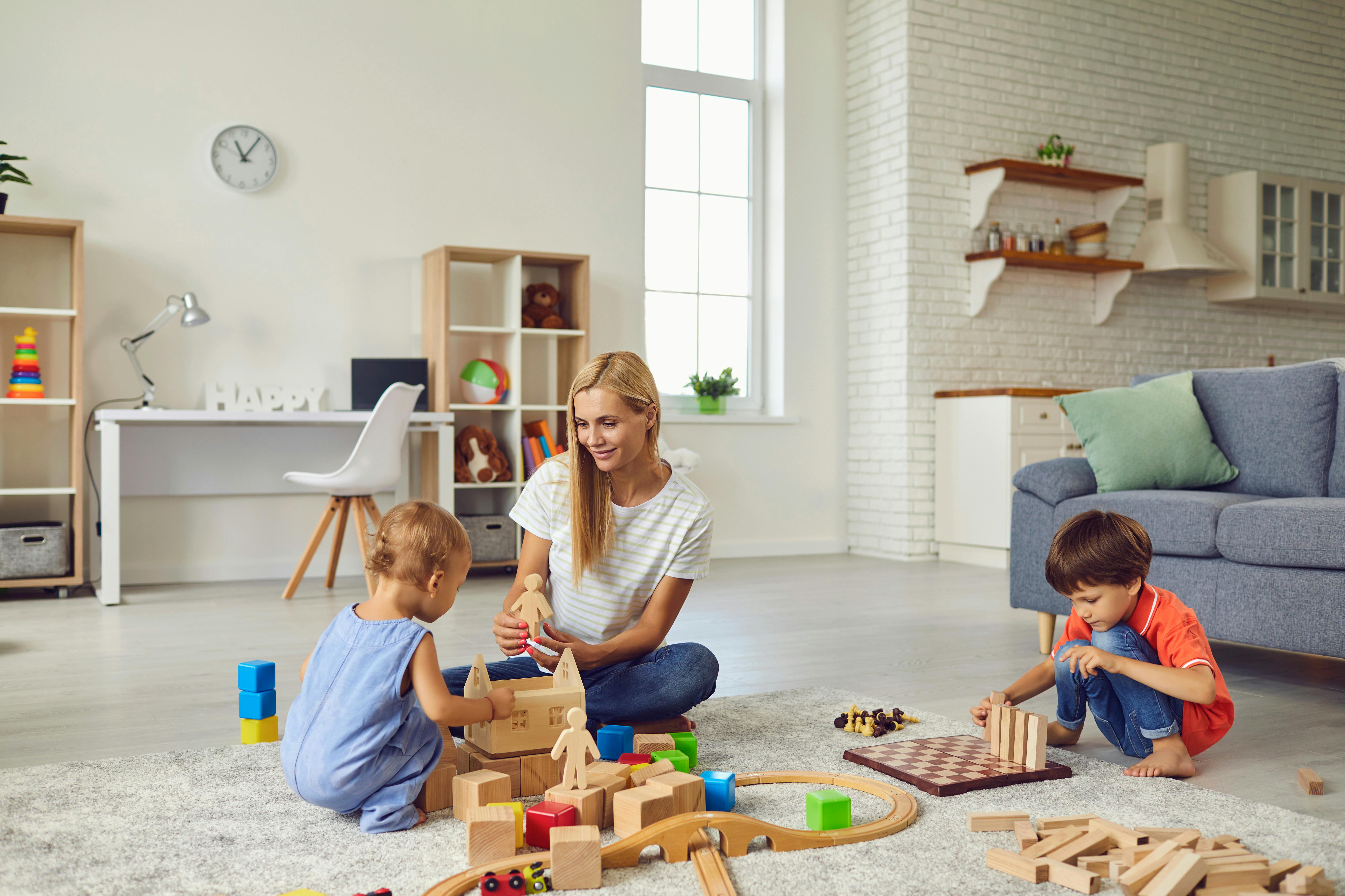 Une femme blonde joue avec deux enfants sur le sol d'un salon.