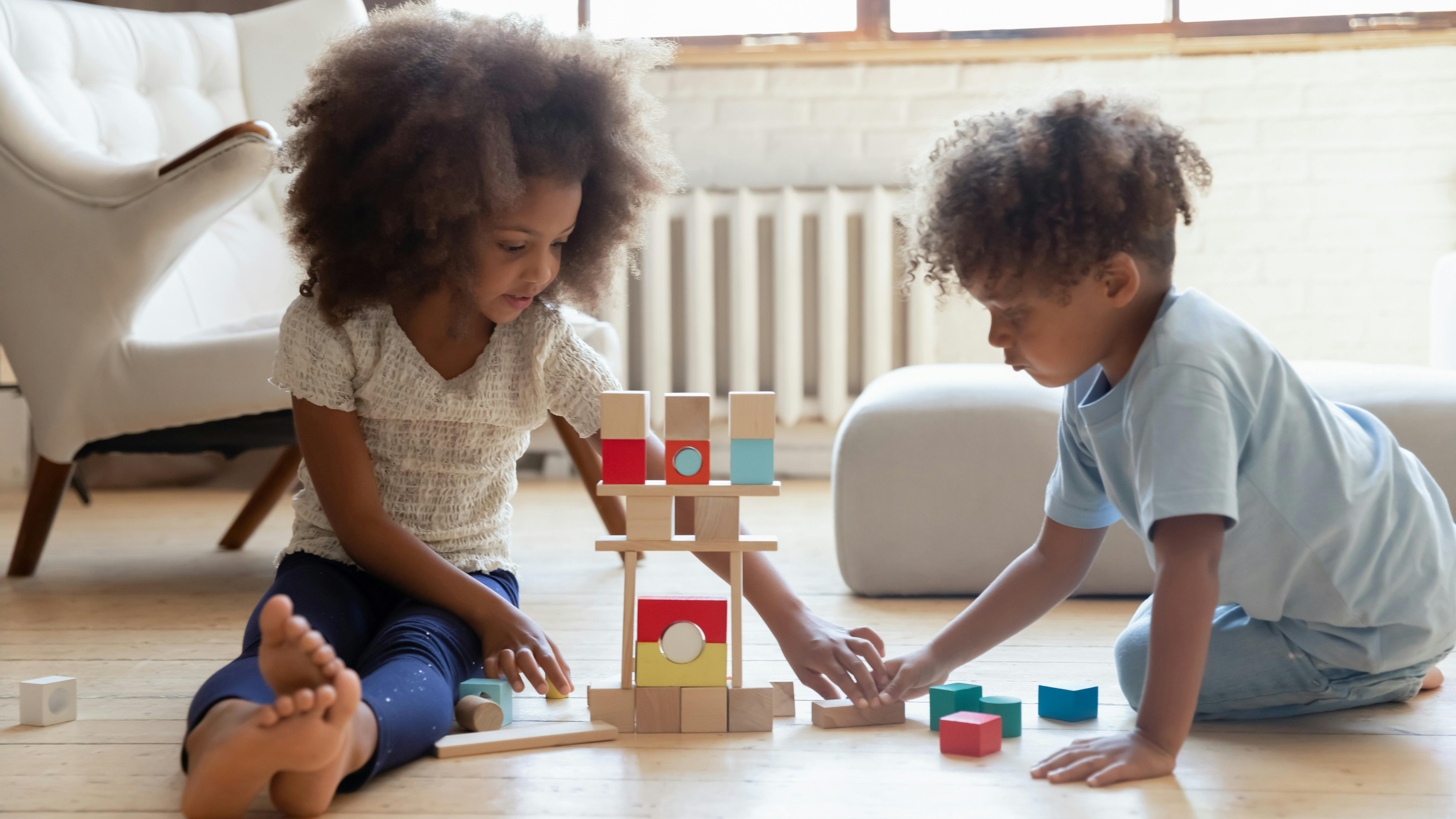 Deux enfants jouent avec des blocs en bois sur le sol d'une pièce ensoleillée.