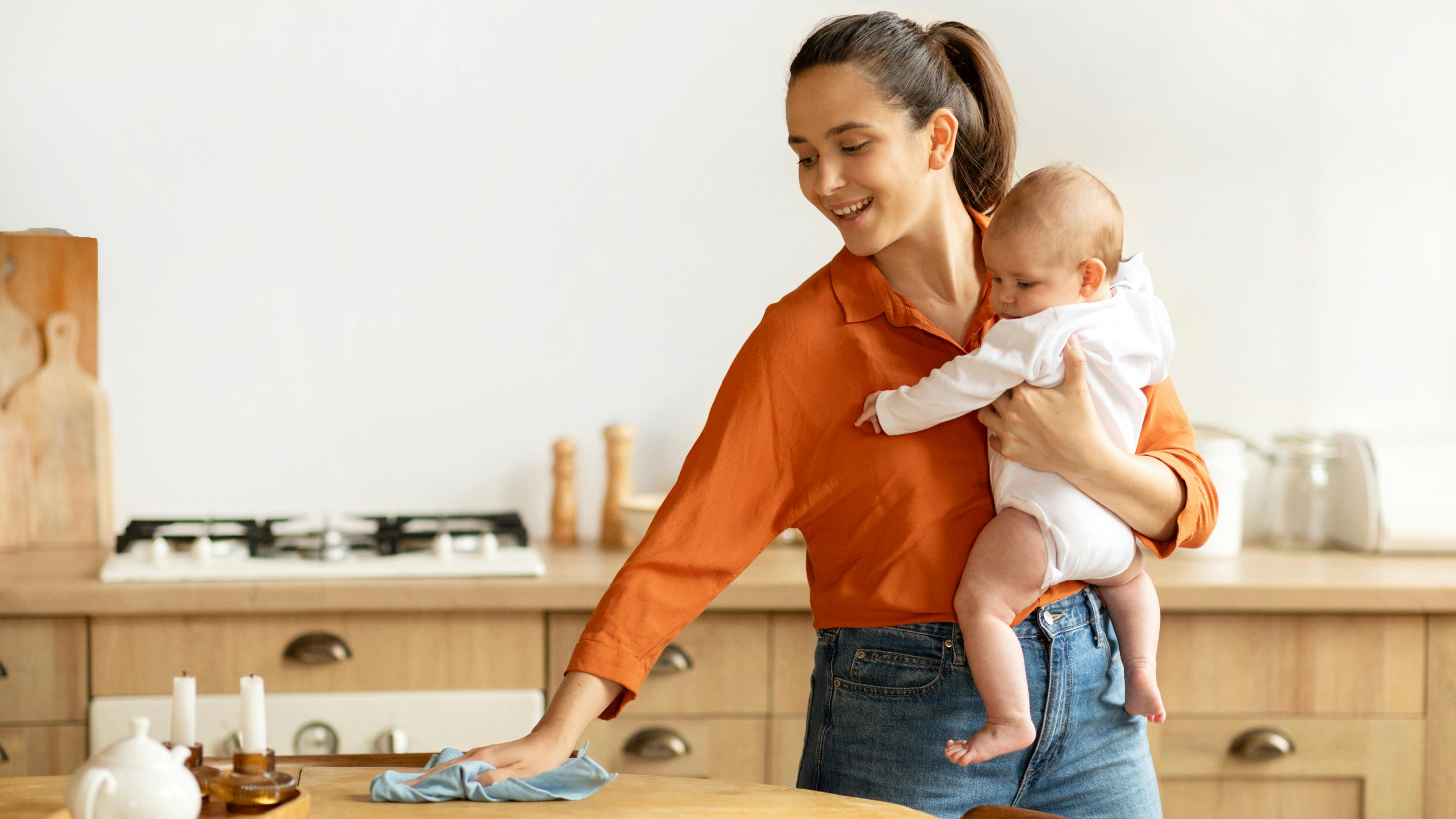 Une femme essuie une table de cuisine tout en tenant un bébé dans ses bras.