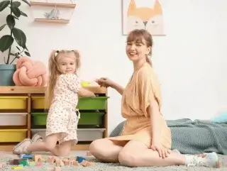 Une jeune femme souriante et une petite fille jouent ensemble sur un tapis dans une chambre d'enfant lumineuse, entourées de cubes en bois.