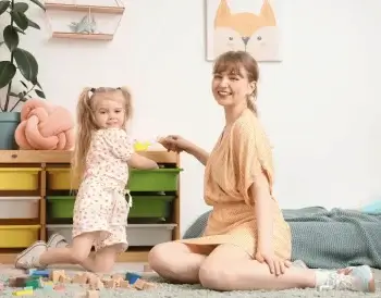 Une jeune femme souriante et une petite fille jouent ensemble sur un tapis dans une chambre d'enfant lumineuse, entourées de cubes en bois.