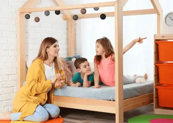 Une assistante maternelle assise à côté d'un lit en bois où se trouvent un petit garçon et une petite fille qui pointe du doigt.