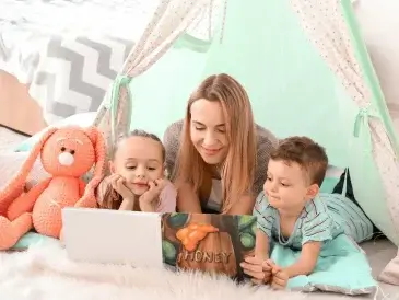 Une nounou lisant un livre avec un petit garçon et une petite fille à l'intérieur d'une tente tipi vert clair.